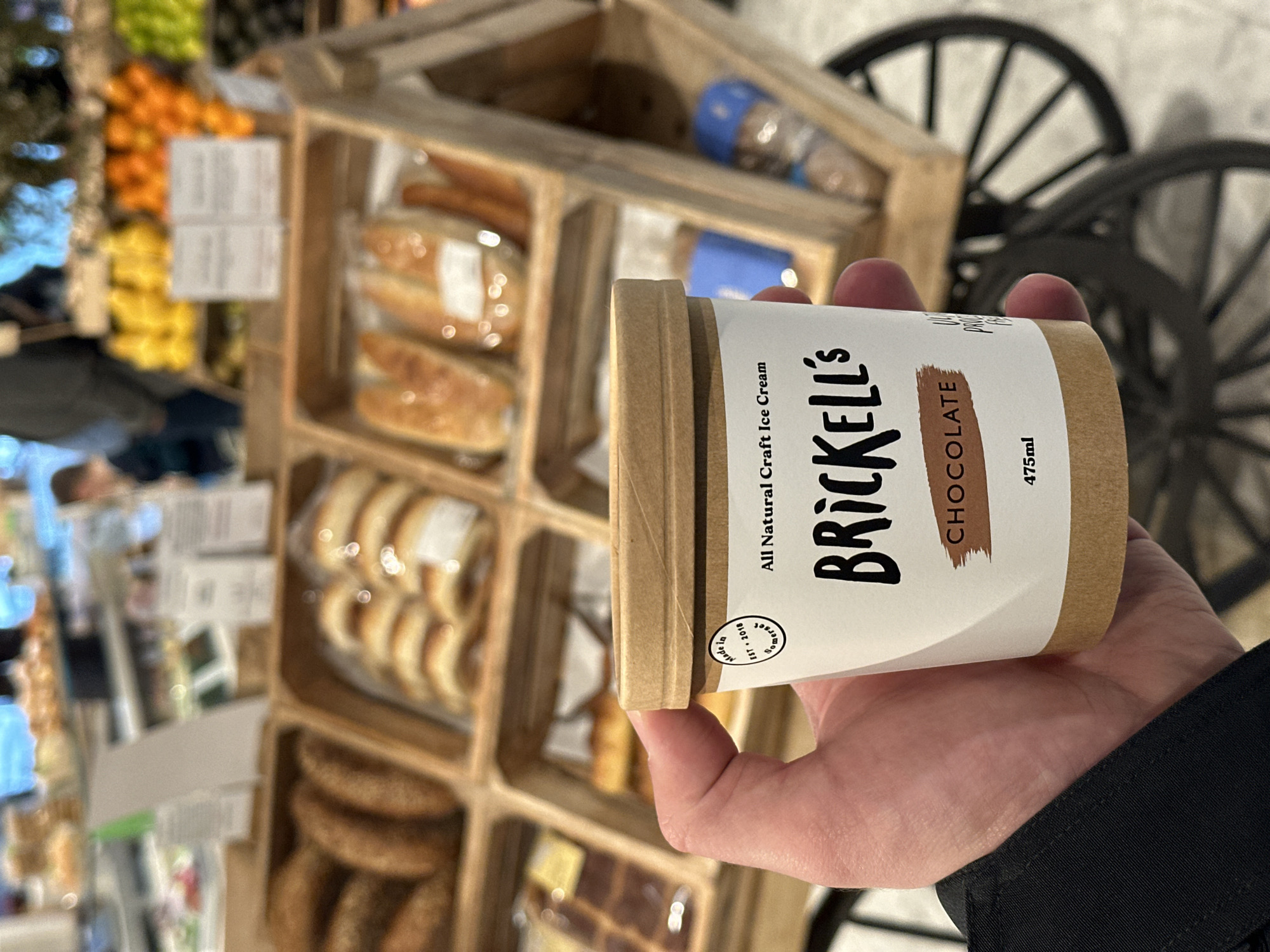 The scene depicts a hand holding a container of Brickell's All Natural Craft Ice Cream, Chocolate flavor, with a label indicating 475ml. The background features a wooden display case stocked with various food items, including what appears to be pastries and breads. In the blurred background, glimpses of fresh produce such as citrus fruits and greenery can be seen, suggesting a market setting. Additionally, a decorative wheel is partially visible behind the display case. The location is possibly in London, United Kingdom, indicated by information on the product label. The focus is on the ice cream container held in hand, with a market environment providing context.