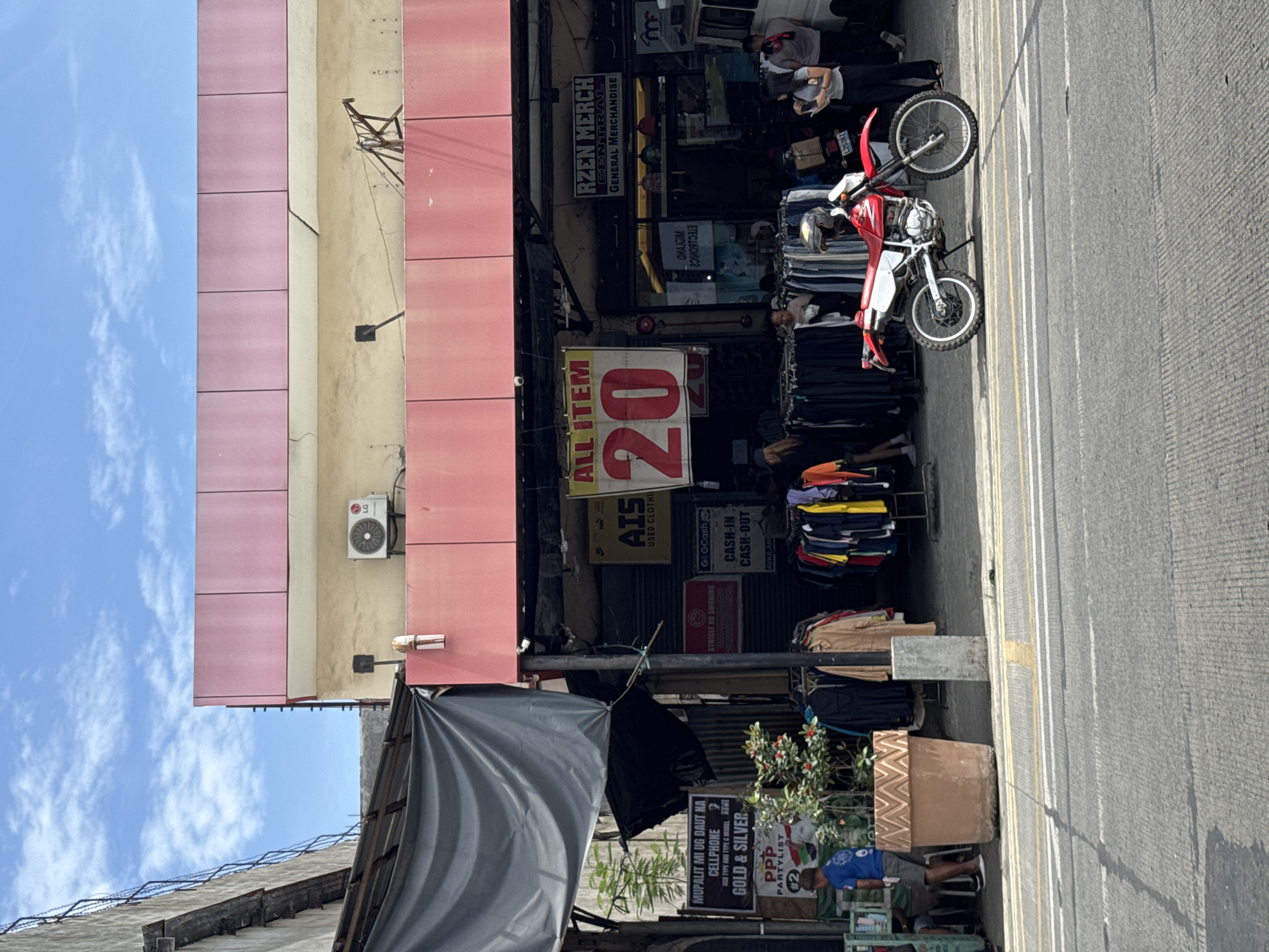 This photograph captures a vibrant daytime street scene in Davao City, Philippines, under a clear blue sky with light clouds.
The left side of the image is dominated by a multi-story building featuring a facade of alternating pink and beige panels. An LG outdoor air conditioner unit is mounted on its upper section. Below, at street level, are various open-front shops and stalls. One prominent sign reads "RZEN MERCH CENTRAL GENERAL MERCHANDISE." Another large, red sign declares "ALL ITEM 20," suggesting a pricing strategy. Adjacent to this, "AIS USED CLOTHI" (likely "Used Clothing") is advertised, with racks of garments displayed outside. Further along, a sign for "G GCash CASH-IN CASH-OUT AVAILABLE" is visible, indicating financial services. A "STRICLY NO SMOKING" notice is also posted.
On the right side of the frame, an asphalt road with yellow and white markings runs parallel to the shops. A red and white dirt bike or enduro motorcycle, with the number "L20382" visible, is parked near the roadside. Several people are present, including one individual seated on the motorcycle and others near the shops, one seemingly occupied with a phone. Further down, under a dark tarpaulin awning, a stall advertises in a local language (Cebuano/Visayan): "MUPALIT MI UG DAUT NA CELLPHONE USO TYPE AND TYPE C MODEL GOLD & SILVER," meaning "We buy broken cellphones of any type and model, gold & silver." A large, decorative planter with a small plant sits nearby, and a person in a blue shirt is seated next to it. Political signage for "PPP PARTYLIST #2" can also be seen. The scene depicts a bustling commercial area, likely a market street, teeming with activity.