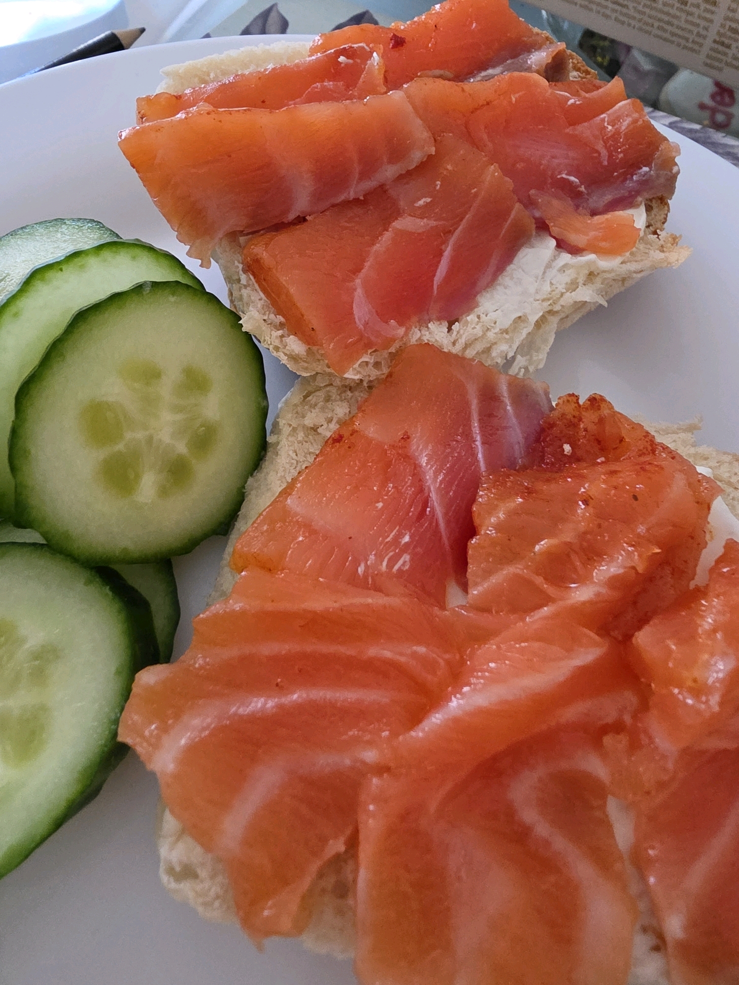 The image shows two open-faced sandwiches with smoked salmon and cream cheese, served with sliced cucumber. The sandwiches are on a white plate. The salmon is thinly sliced and arranged on top of the cream cheese. The cucumber slices are circular and have a light green skin and a pale green interior with visible seeds. The background is blurred but suggests an indoor setting with a table and possibly some patterned fabric. There is no indication of the specific location in West Suffolk, United Kingdom. The scene appears to be a close-up shot of food, likely prepared for a meal.
