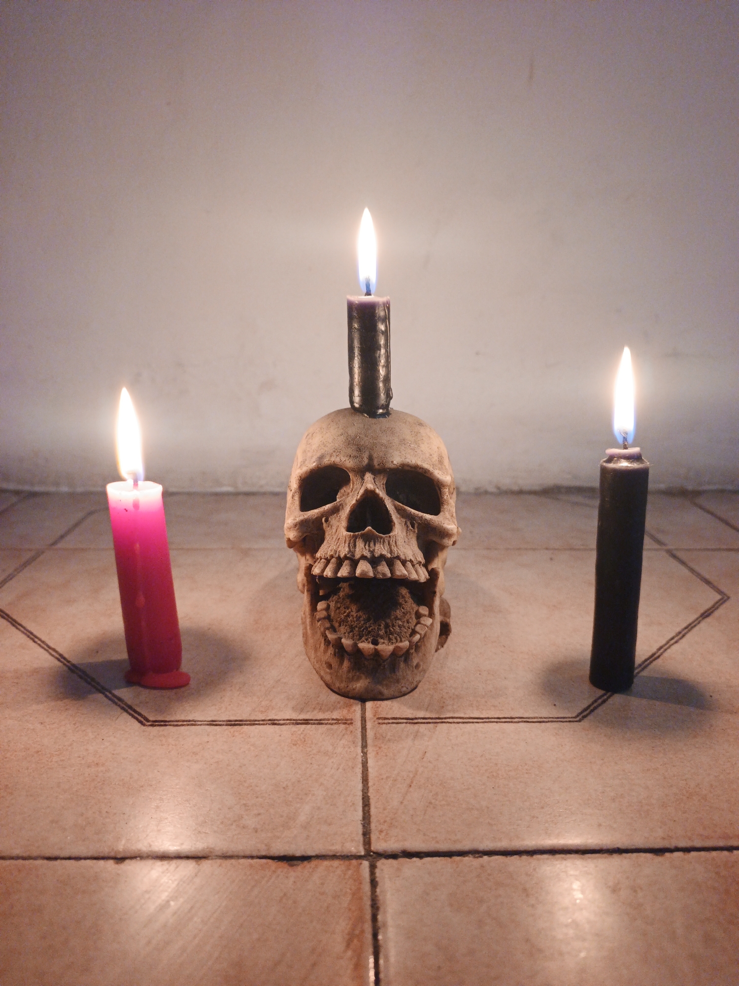 The image shows a skull with a candle on top, flanked by a pink candle on the left and a black candle on the right. All three candles are lit. The background is a plain wall. The skull and candles are placed on a tiled floor.
Based on the location information provided, it can be assumed that this image was taken in Tinaquillo, Venezuela. However, the image itself provides no further information about the specific location within the city.