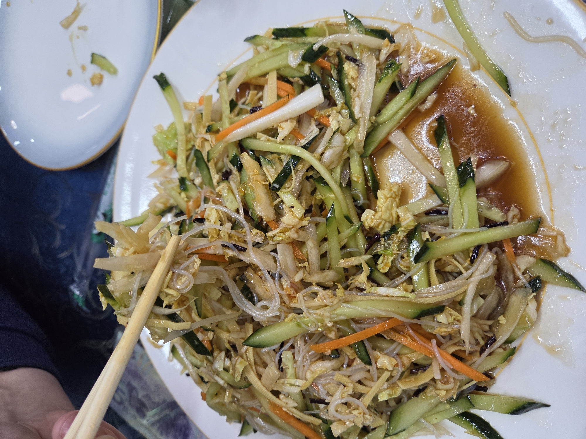 An overhead shot captures a close-up of an Asian-style cold dish or salad served on a large, white round plate with a subtle gold rim. The plate is filled with a colorful medley of finely cut vegetables and translucent noodles, coated in a glossy, brown sauce that collects at the bottom.
The main ingredients visible are julienned bright green cucumbers, thin orange carrot strips, and delicate, almost transparent glass noodles. Also present are pale, shredded vegetables, possibly cabbage or bean sprouts, and thin, light-colored strips that could be bamboo shoots or tofu skin. Darker flecks, possibly black fungus or sesame seeds, are interspersed throughout the dish.
In the lower-left corner, a person's hand, wearing a dark blue garment, holds a pair of light wooden chopsticks. The chopsticks are positioned over the food, suggesting the act of eating or serving.
To the upper-left, partially out of focus, is another white, oval plate with a similar gold rim, appearing mostly empty except for a few small food scraps and sauce residue. The lighting suggests an indoor setting, likely a restaurant or home dining table. No text is visible in the image.