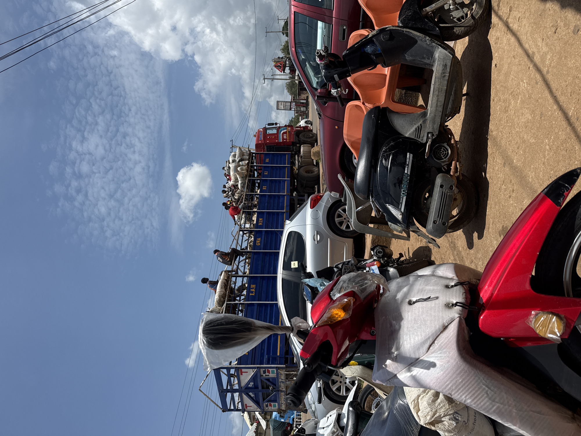 This is a full-shot, eye-level, outdoor photograph taken in the daytime. In the foreground, a red and white scooter is parked and covered in protective plastic sheeting. To the right, a black scooter with orange plastic seats is also parked. In the middle ground, a silver car is parked behind the scooters. Further back, a large blue truck with people and cargo on top is parked. The truck is decorated with writing that includes "LAH GWA NIN". A red truck is parked to the right of the blue truck. The environment appears to be a busy street with some buildings and power lines visible in the background. The sky is partly cloudy.