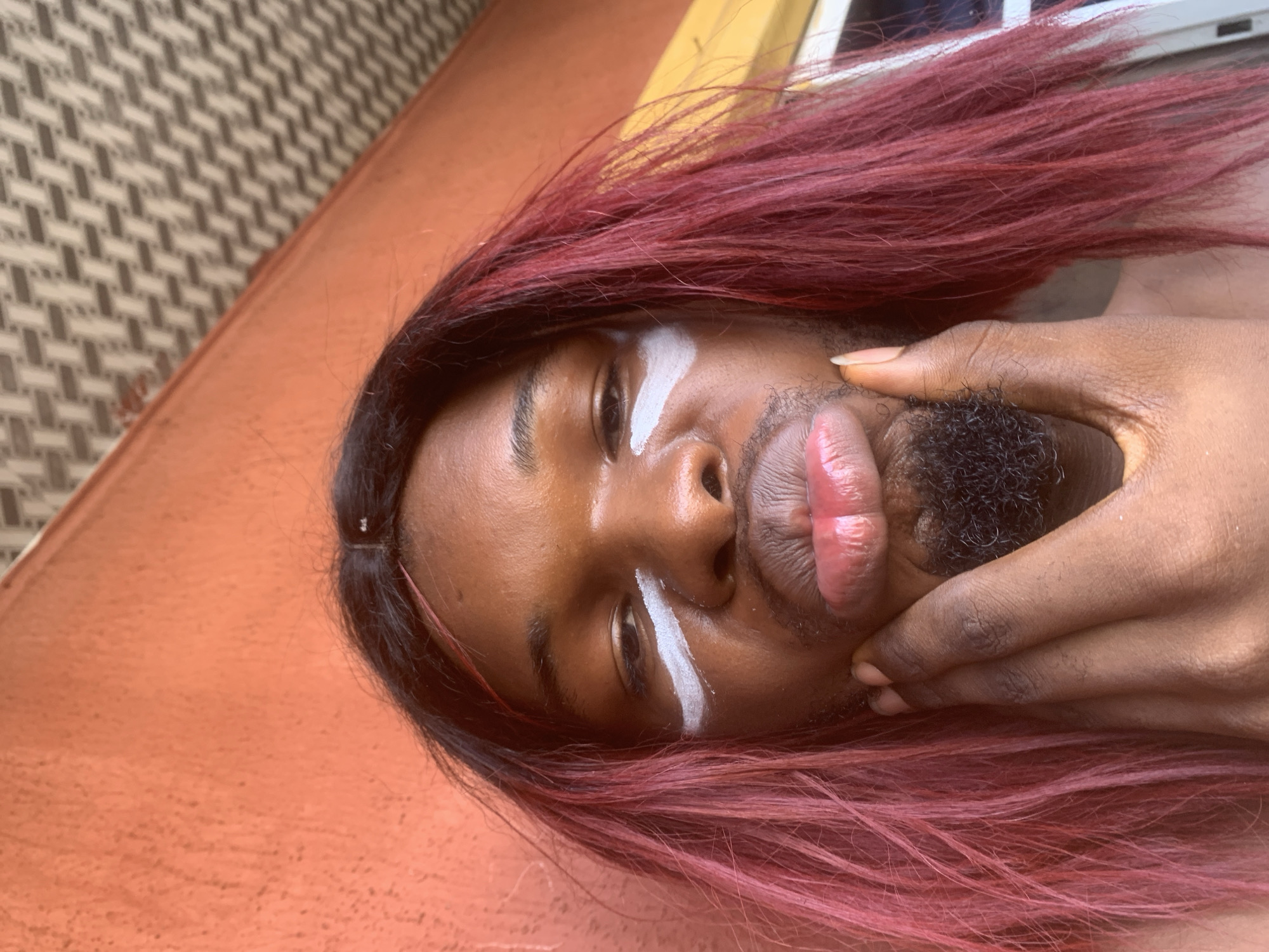 A person with dark skin and long, reddish-pink hair is prominently featured in a close-up, angled shot. The individual's head is tilted sideways, with their gaze directed towards the camera. White, artistic stripes are painted under each eye, and their lips are full, appearing glossy or well-moisturized, and slightly puckered. There is visible dark facial hair around the mouth and chin area.
One hand, with dark skin, is visible on the right side of the frame, gently holding or squeezing the person's lower face, near the chin and mouth. The skin has a luminous sheen, suggesting natural light and perhaps moisturized skin.
The background consists of two distinct areas: an orange-brown or terracotta-colored surface that takes up a significant portion of the lower and right side of the image, and a patterned, woven-like surface in the upper left corner. The lighting appears to be natural daylight, highlighting features on the face. There is no visible text in the image.