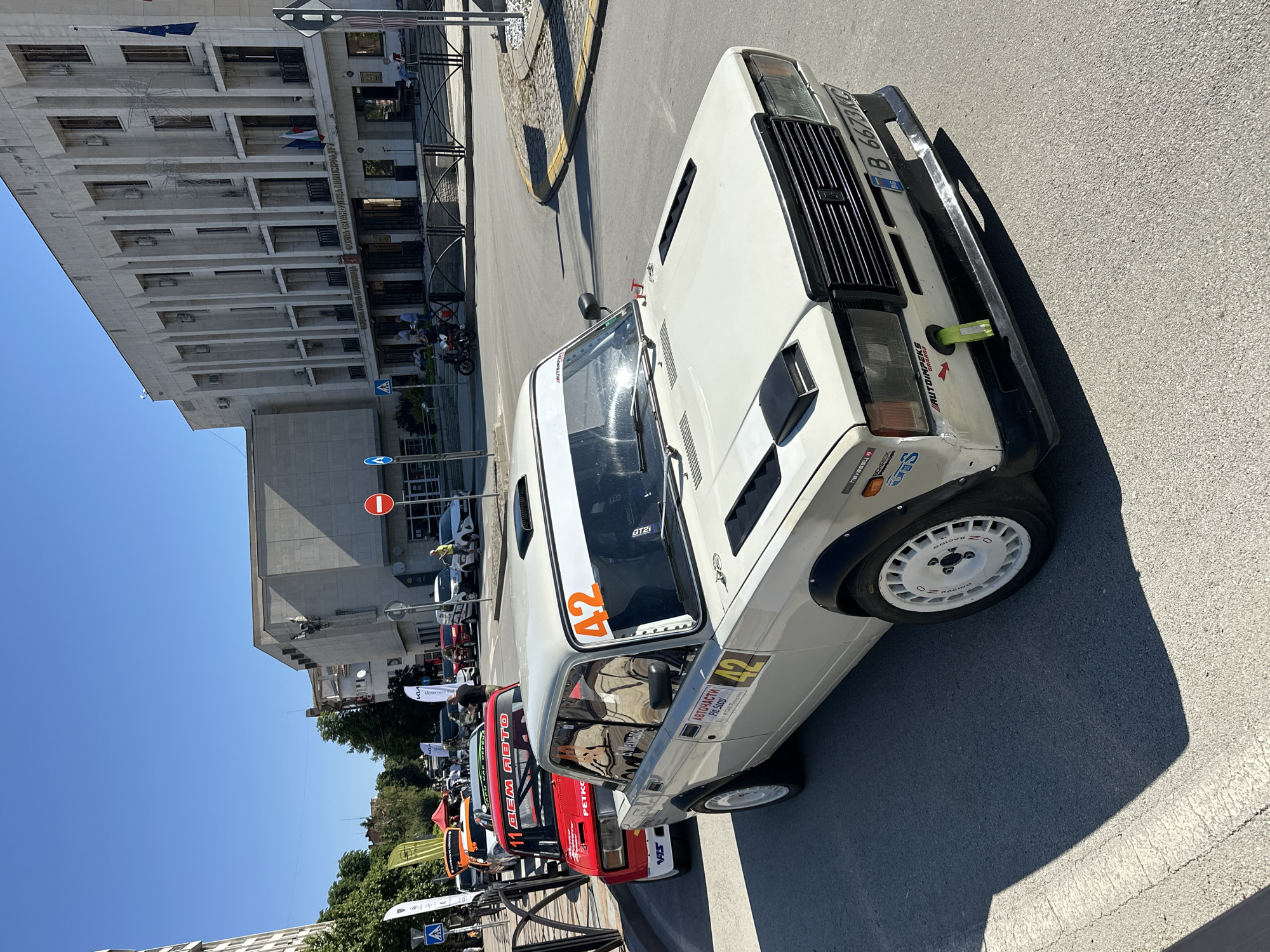 In the city of Gorna Oryahovitsa, Bulgaria, a lineup of rally cars is parked on a paved street in front of a large, ornate building with a concrete facade. The building displays Bulgarian and EU flags and has the inscription "Gorna Oryahovitsa Municipality". The street is lined with trees and other buildings in the background. Several people are visible, some standing near the cars and others walking on the sidewalk. A few motorcycles are also parked in front of the building. The main focus is on a white rally car in the foreground, numbered '42', with modifications for racing, such as a roll cage, racing tires, and aerodynamic enhancements. Other rally cars, predominantly red, are parked behind it. The scene appears to be a gathering or an event related to automotive sports.