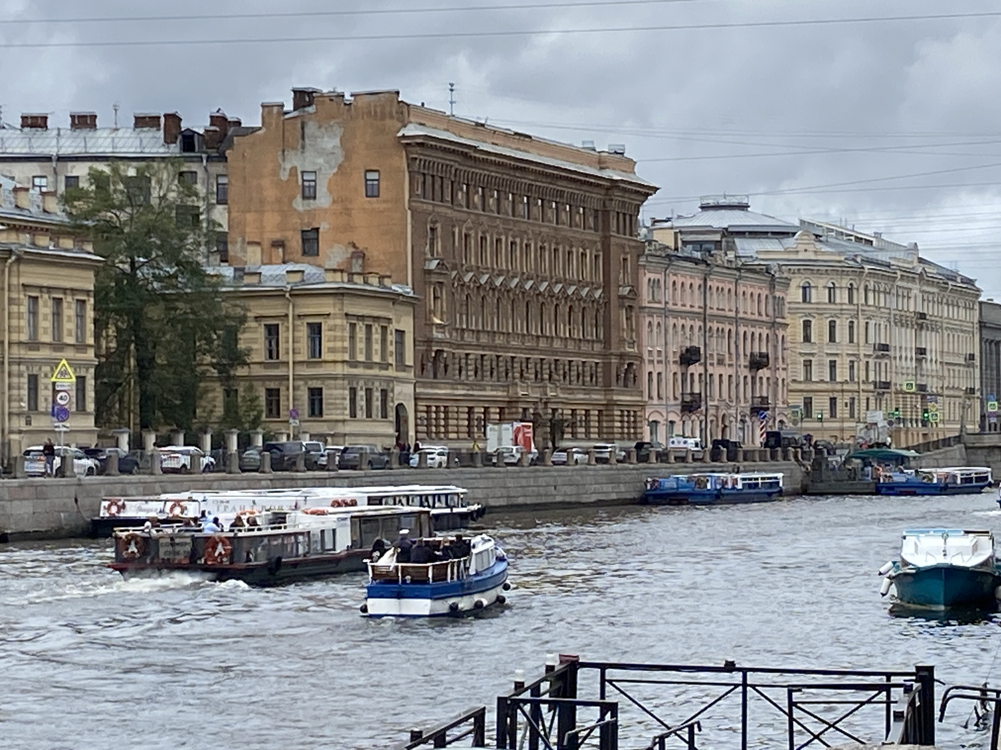This image depicts a cloudy day in Saint Petersburg, Russia, likely along the Neva River or one of its canals. In the foreground, a body of water is visible, with several boats on it. A small blue and white boat with several passengers is moving towards the right. To the left, a larger white boat with a dark hull and passengers is docked or moving slowly. In the middle ground, a stone embankment lines the water, with a row of parked cars and a few pedestrians visible. Behind the embankment, a series of multi-story buildings with ornate architecture and varied facades are prominent. These buildings are characteristic of Saint Petersburg's historic center, with features such as mansard roofs, arched windows, and decorative cornices. The sky is overcast, contributing to a muted color palette. The overall scene suggests a typical urban landscape with both transportation on the water and architectural grandeur on land.