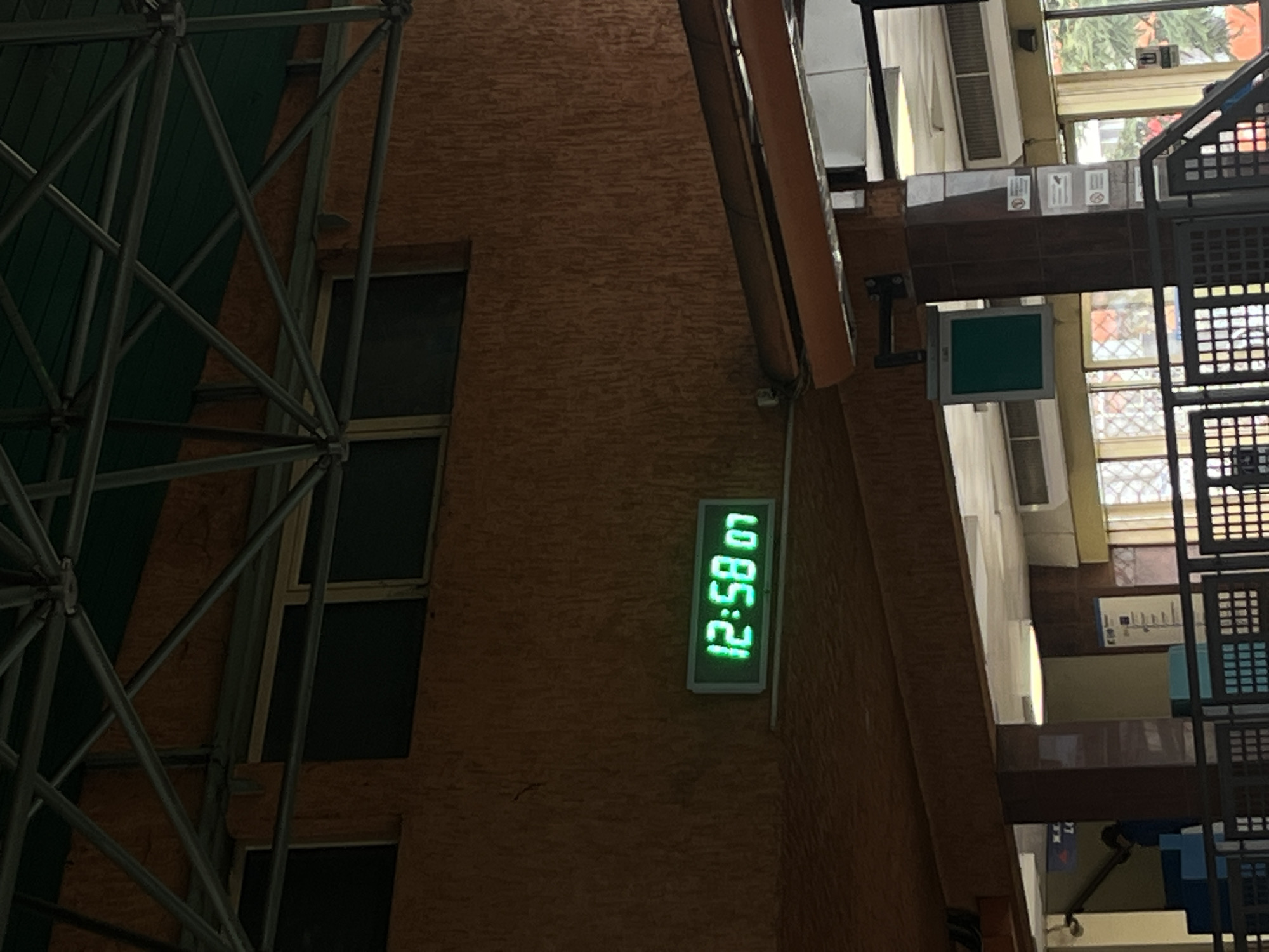 The image shows a view of an indoor space with architectural elements. On the left side, a metallic structure with diagonal supports is visible. Adjacent to it is a textured brick wall with two dark, rectangular windows. On the wall, a digital clock displays "12:58 07" in green digits. To the right, there is a multi-story structure with window openings and metal grating. The scene appears to be inside a building with natural light entering from the upper windows. Based on the presence of a digital clock, and architectural design, it is possible it is in a public building in the city of Sofia, Bulgaria, however, further detail of the surroundings are needed to be sure.