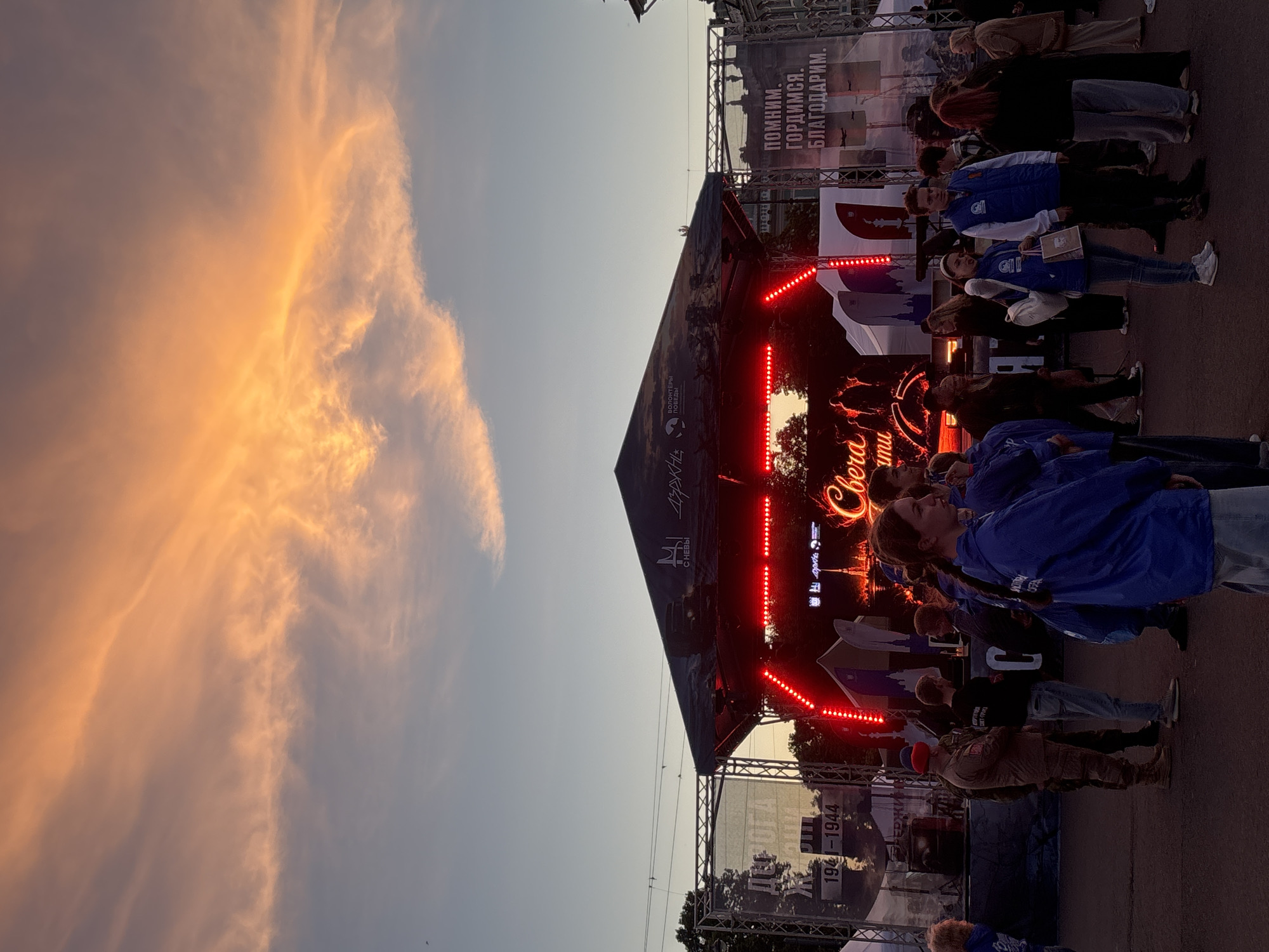 The image depicts an outdoor gathering in Saint Petersburg, Russia, near a stage set up for an event. The sky is painted with vibrant orange and pink clouds, indicative of a sunset. A stage is visible, adorned with red LED lights and a large screen displaying text and imagery, including the words "Свега" (meaning "Light" or "Snow") in a prominent, stylized font. Two large banners are present on either side of the stage. The left banner features the text "ДОРОГА" (meaning "Road") and the dates "1941-1944". The right banner reads "ПОМНИМ. ГОРДИМСЯ. БЛАГОДАРИМ." (meaning "We Remember. We Are Proud. We Thank."). A crowd of people, many wearing blue vests with logos, are gathered in front of the stage. Some individuals appear to be volunteers, holding bags or participating in the event. The overall scene suggests a commemorative event, possibly related to Victory Day or a similar historical occasion, taking place in a public square or open area in the city.