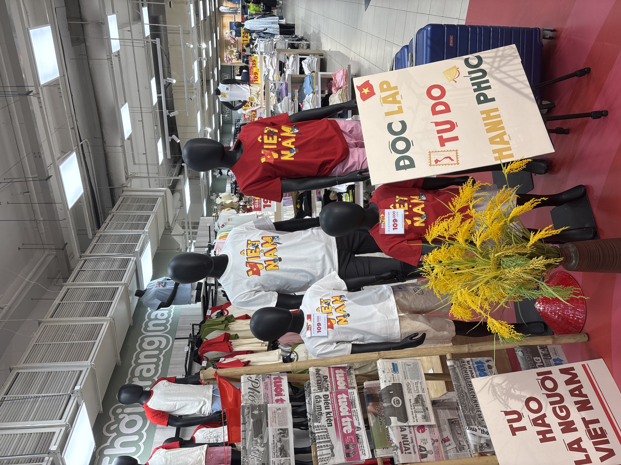 The image shows a display of T-shirts with "VIỆT NAM" (Vietnam) printed on them. They are on mannequins with black heads in what appears to be a store. Signage reads "ĐỘC LẬP TỰ DO HẠNH PHÚC" (Independence, Freedom, Happiness) and "TỰ HÀO LÀ NGƯỜI VIỆT NAM" (Proud to be Vietnamese). Newspaper clippings are also part of the display. The location is most likely a department store or similar retail environment. The location in the city of Đà Nẵng, Vietnam is not indicated.