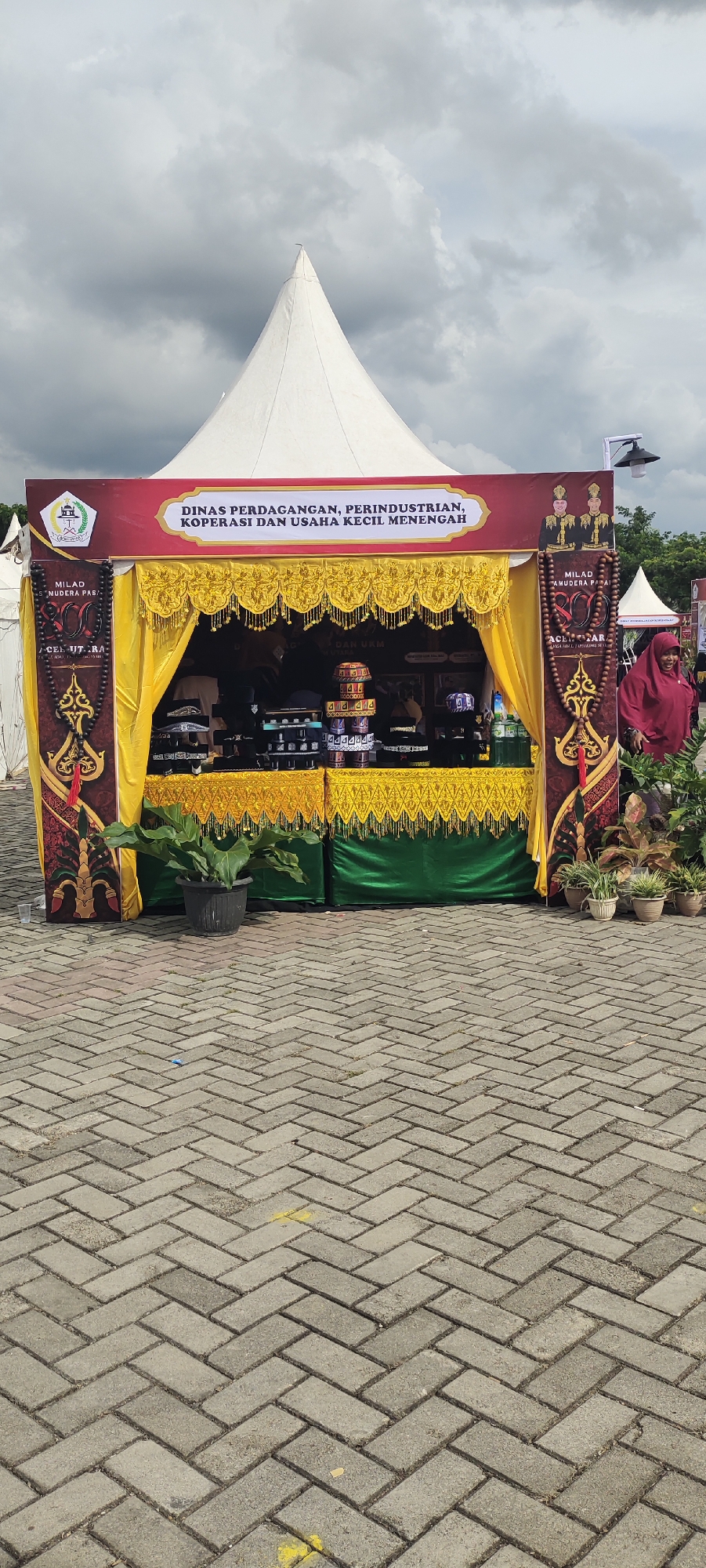 An outdoor event is taking place under a cloudy sky. A white, conical tent is the dominant structure, with a maroon and gold stall set up in front of it. The stall has a sign that reads "DINAS PERDAGANGAN, PERINDUSTRIAN, KOPERASI DAN USAHA KECIL MENENGAH" which translates to "Department of Trade, Industry, Cooperatives, and Small and Medium Enterprises." The stall displays various items, including hats, beaded necklaces, and bottled liquids. Two individuals in traditional Indonesian attire are depicted on the stall's signage.
To the right of the stall, a woman wearing a red headscarf and a dark red shirt is visible, smiling and looking towards the viewer. A few potted plants are placed around the stall and to the right of the woman. The ground is paved with interlocking gray bricks. The overall scene suggests a public exhibition or market in Meunasahaluedrienl Hoksukon Barat, Indonesia.