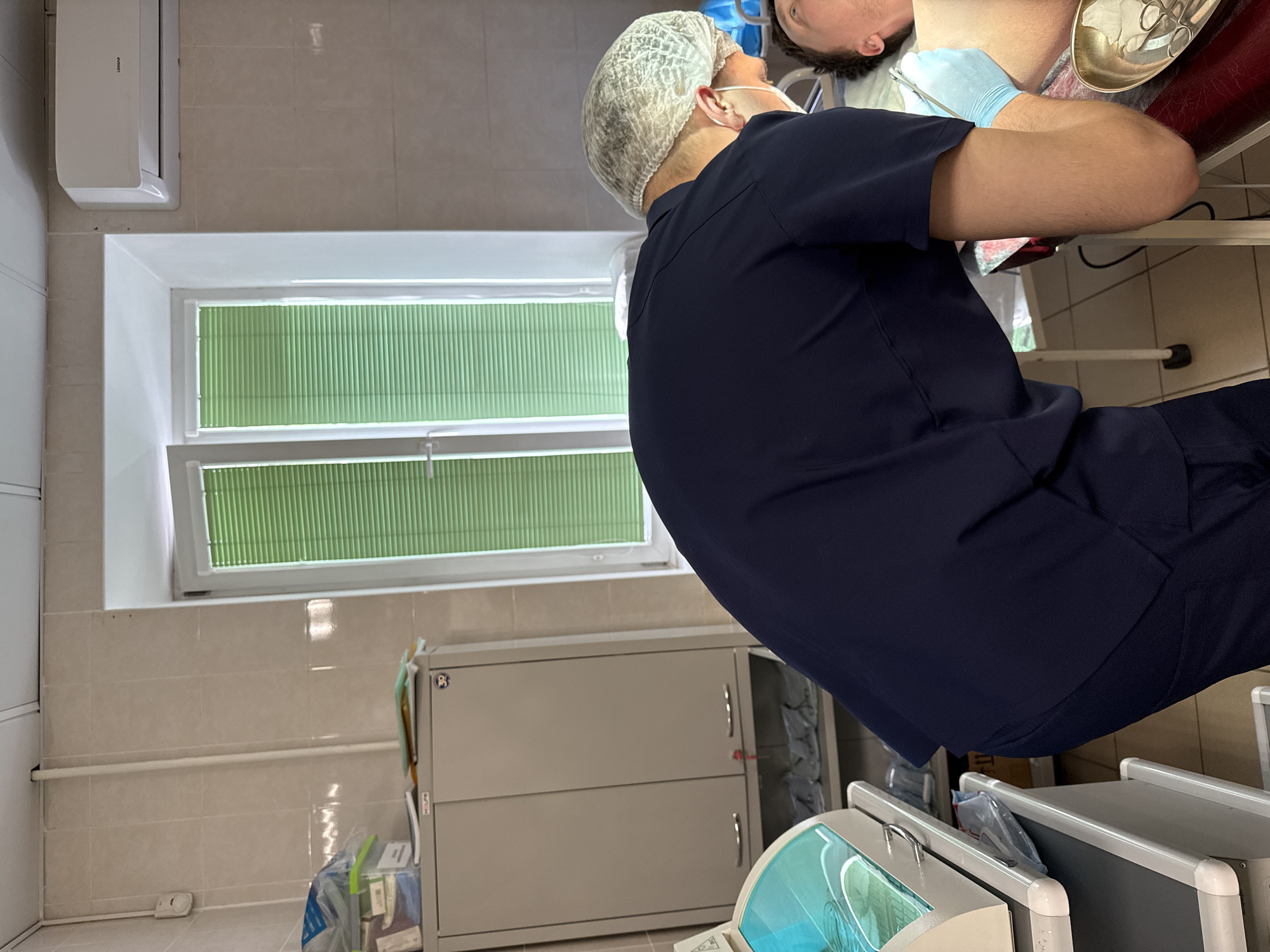 In a brightly lit room in Perm, Russia, a medical professional wearing a dark blue scrub suit and a hairnet is examining a patient. The patient is lying on their back, with a blue surgical drape covering their upper chest. A metal tray containing surgical instruments is positioned near the patient's head. The room appears to be a clinic or examination room, with a window covered by green blinds. A metal storage cabinet stands against the wall, near a dental X-ray machine. An air conditioning unit is affixed to the upper wall.