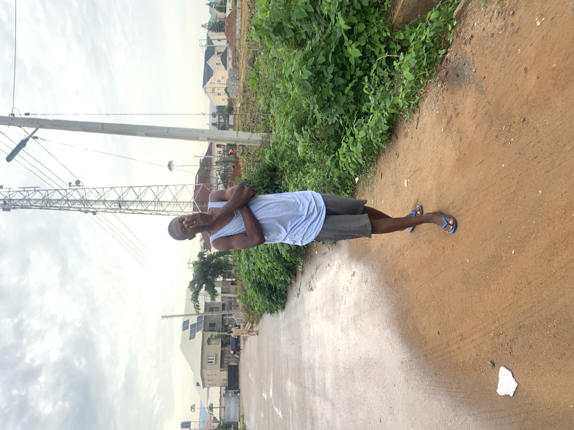 A young man stands on a dirt path in Abuja, Nigeria, with his hands covering his mouth and nose. He is wearing a light blue tank top and grey shorts, with blue flip-flops on his feet. To his left, a wet, paved road curves away, with several buildings visible in the background, some featuring solar panels. A large telecommunications tower and utility poles stand behind him. The area is bordered by lush green vegetation and appears to be under construction, with piles of sand and building materials present. The sky is overcast, suggesting a cloudy day.