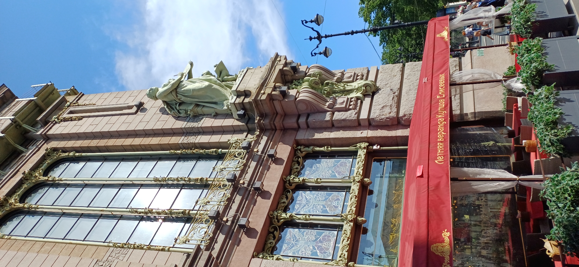 This media file captures a low-angle view of the exterior of a historic building in Saint Petersburg, Russia, on a bright, sunny day. The sky above is a clear blue with scattered white clouds. The building features ornate architecture, including intricate sculptures and detailed window frames.
Prominently displayed on the facade is a large, pale green statue of a reclining figure holding a scroll and a pen, suggesting artistic or scholarly pursuits. Adjacent to the statue, green, stylized sculptural elements adorn the stone facade. Below, large, arched windows with decorative golden trim are visible. Some of these windows feature stained glass with intricate patterns in shades of blue and purple.
A vibrant red banner hangs vertically along the right side of the building. This banner has text in Russian written in a lighter color, which appears to read "Летняя веранда Купцов Елисеевых" (Summer Veranda of the Merchants Eliseevs). The banner is adorned with a golden, stylized illustration of an animal, possibly a bird or a griffin, at its bottom. Artificial greenery cascades down the right edge of the building, adding a touch of nature to the urban setting. A street lamp with two lights is also visible against the sky.
In the background, through some of the upper windows, several people can be seen moving about, indicating that the building is likely a commercial establishment or a public space that is open. The overall impression is one of grandeur and historical significance, with the bright sunlight highlighting the detailed craftsmanship of the building.