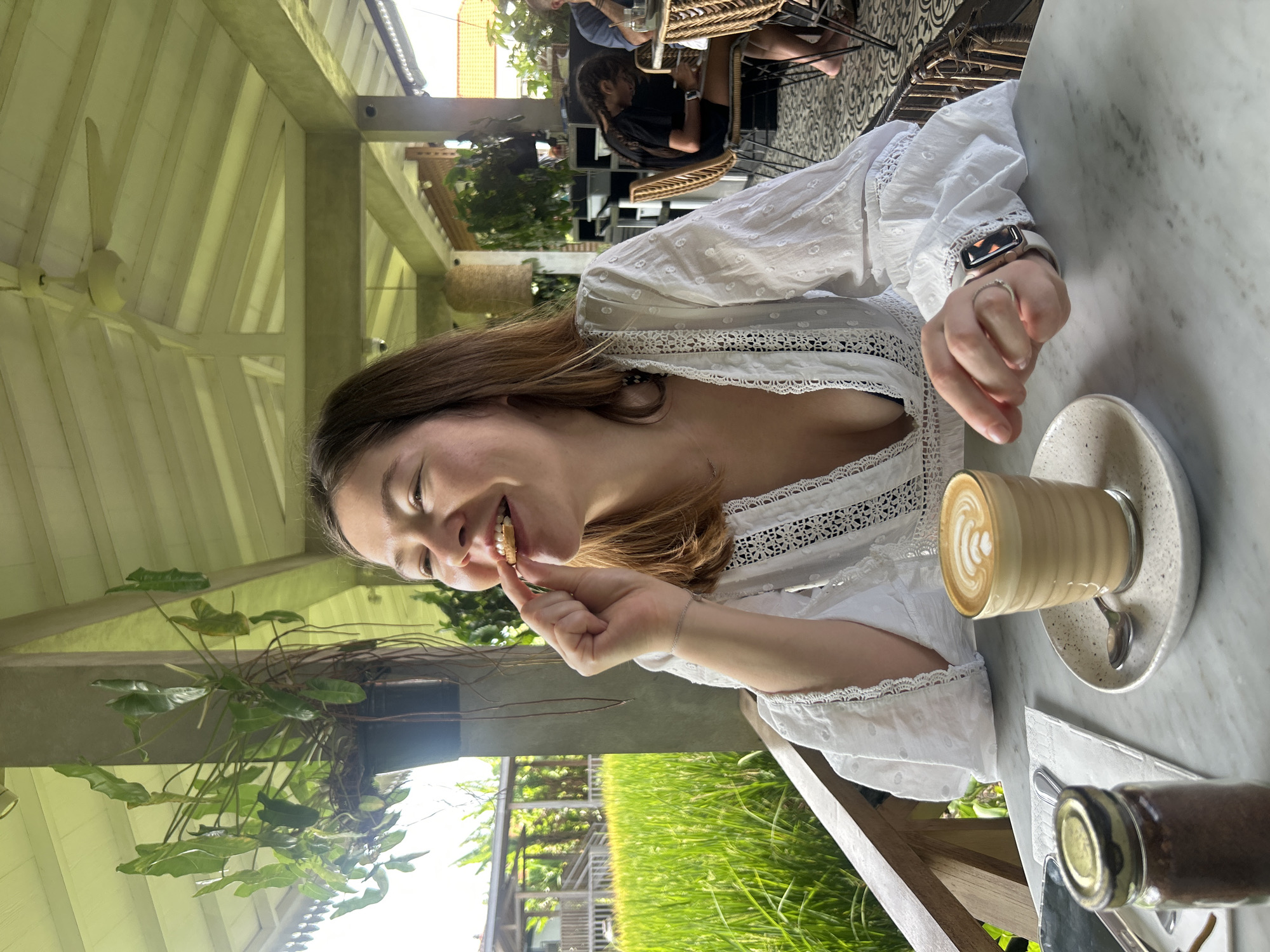 A young woman with brown hair is shown from the chest up, seated at a marble table with a cup of coffee and a small jar of what appears to be coffee grounds. She is wearing a white eyelet blouse and is in the process of eating a cookie, with her eyes closed and a smile on her face. The table is set on a balcony or patio overlooking a lush green rice field. In the background, other patrons can be seen at tables, and a ceiling fan is visible overhead. The scene appears to be set in a cafe or restaurant in North Kuta, Indonesia, known for its scenic rice paddy views.