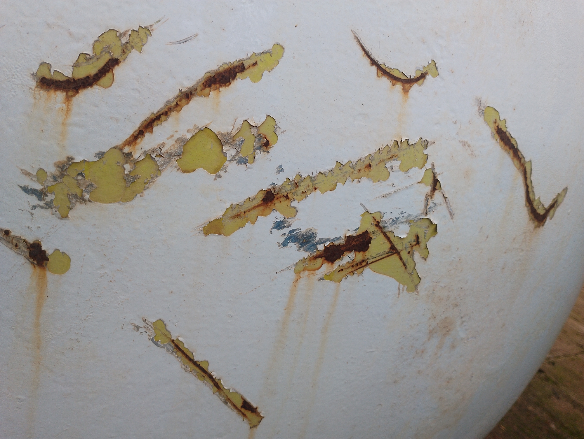 The image shows a close-up, detailed view of peeling white paint with visible rust and underlayer damage. The texture is rough and irregular, with patches of yellow and brown showing through the compromised white surface. Several thin, curved streaks of rust are prominent, indicating areas of corrosion. Some areas exhibit slight blue discoloration beneath the peeling paint. The overall impression is one of wear and decay on a possibly metallic or painted surface. The context of Jos, Nigeria, is not visually discernible from the image itself.