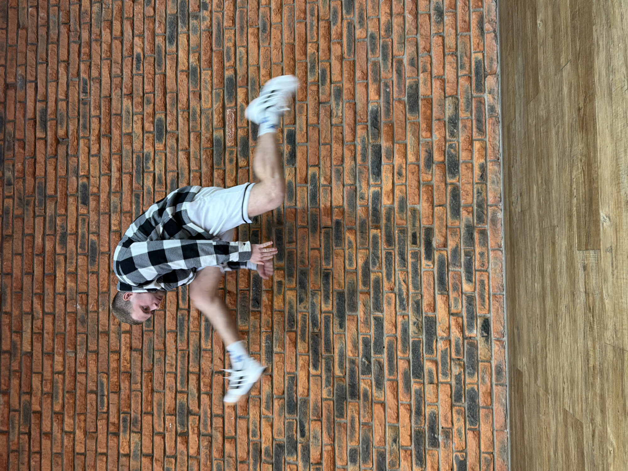 An athletic young man performs an inverted, acrobatic pose against a textured background in Chaikovsky, Russia. The image is rotated 90 degrees clockwise, depicting the man seemingly on a vertical surface. He is dressed in a black and white plaid long-sleeve shirt, white athletic shorts, white socks with blue stripes, and white athletic shoes, which appear slightly blurred, indicating movement. His head is visible, positioned towards the lower left of the frame, with his hands bent near his head. One leg is bent, and the other is extended upwards, creating a dynamic pose.
The background is composed of two distinct materials. The left and larger portion is a brick wall featuring a pattern of reddish-brown bricks mixed with darker, almost black, bricks. To the right of the brick wall is a light to medium brown wooden surface with a visible vertical grain, likely a wooden floor or paneling. The lighting is bright and even, suggesting an indoor environment or an outdoor setting with diffuse light. No specific time of day is discernible. The man's expression is focused, characteristic of an athletic or performance activity. There is no discernible text within the image apart from potential branding on his blurred shoes.