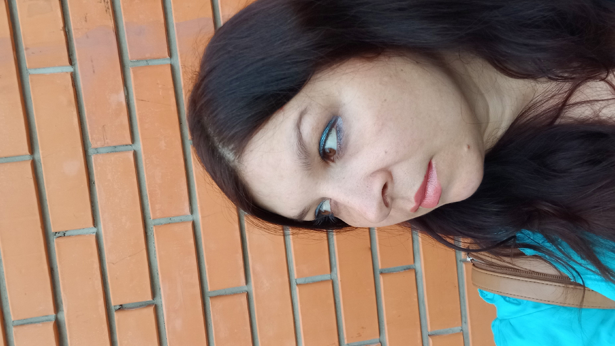 A woman with dark brown hair and blue eyeshadow is standing in front of an orange brick wall. She is wearing a bright blue top and a brown purse strap is visible over her shoulder. She is looking to her left. The image is a medium close-up, taken from the chest up. The brick wall appears to be a facade of a building. The scene is outdoors. There is no discernible context of the city of Kropyvnytskyi, Ukraine.
