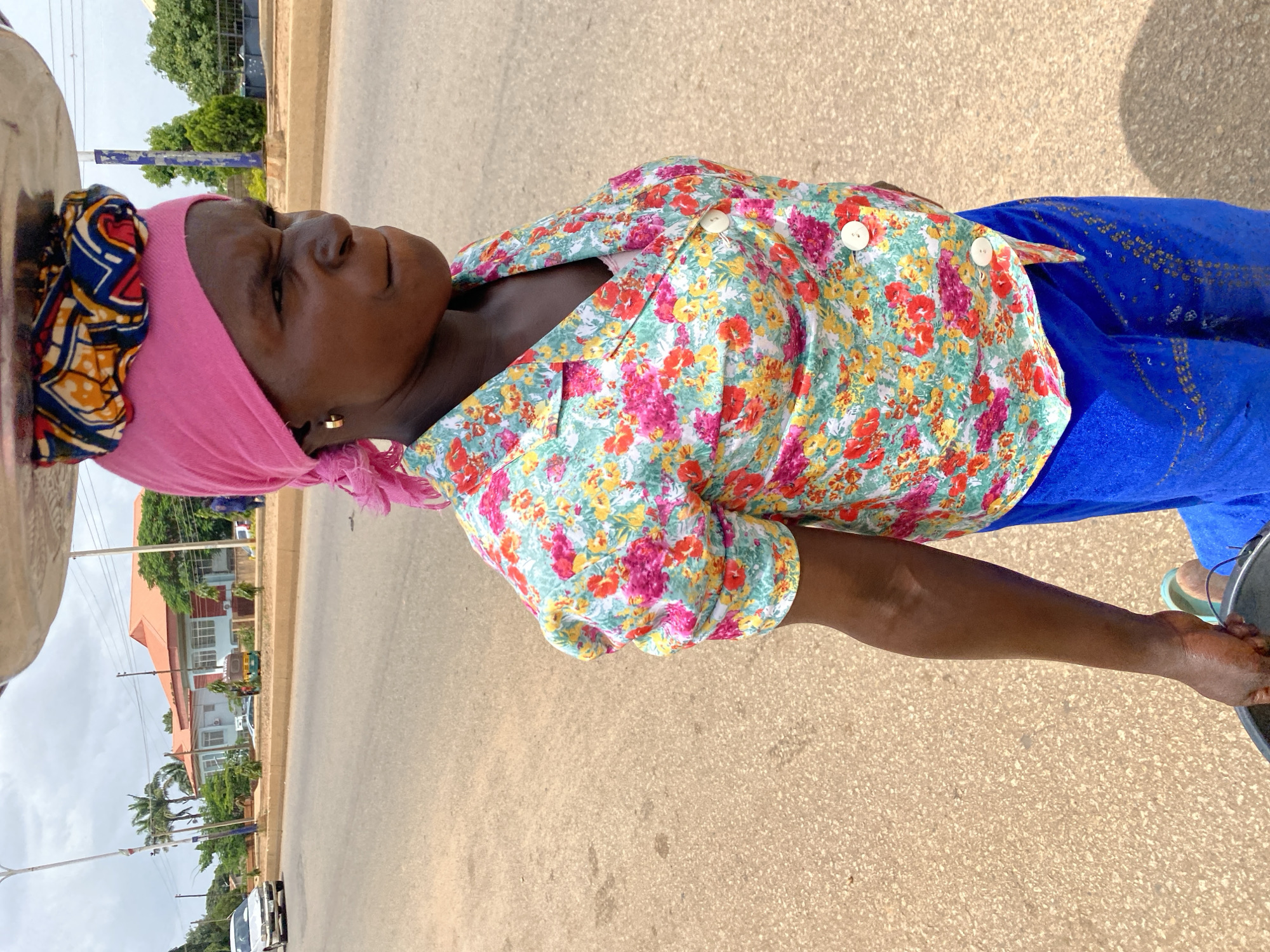 A woman wearing a pink headscarf and a floral-print shirt walks on a paved road in Bukuru, Nigeria. She is balancing a large, flat metal tray on her head, and she carries a black bucket in her right hand. Her expression is stern as she looks to her right. The road is bordered by a concrete median with sparse greenery, and buildings and trees are visible in the background. A white car drives on the road in the distance. The sky is overcast.