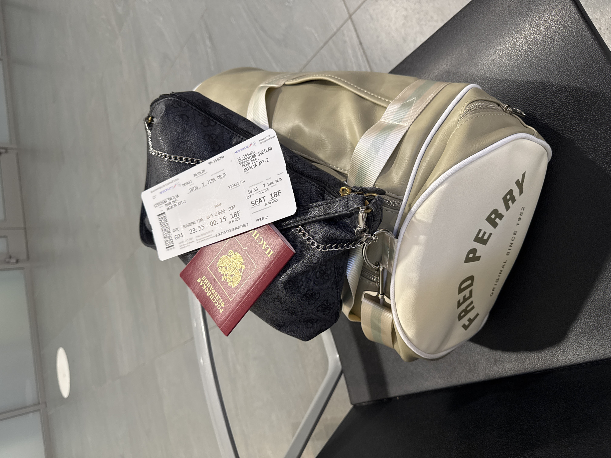 An overhead shot shows a passport and boarding pass resting on top of a black designer bag, which is itself placed on a tan duffel bag with the "FRED PERRY" logo prominently displayed. The scene appears to be at an airport, with a blurred background suggesting waiting area seating and floor tiles. The passport is red and shows the Russian Federation emblem, while the boarding pass indicates a flight from GOSHTINE-SVETLAN to PERERIL, with a departure time of 23:55 on July 31st, 2023, and seat 18F. The location is identified as the city of Bolshoye Savino, Russia, which is likely where the airport is situated.