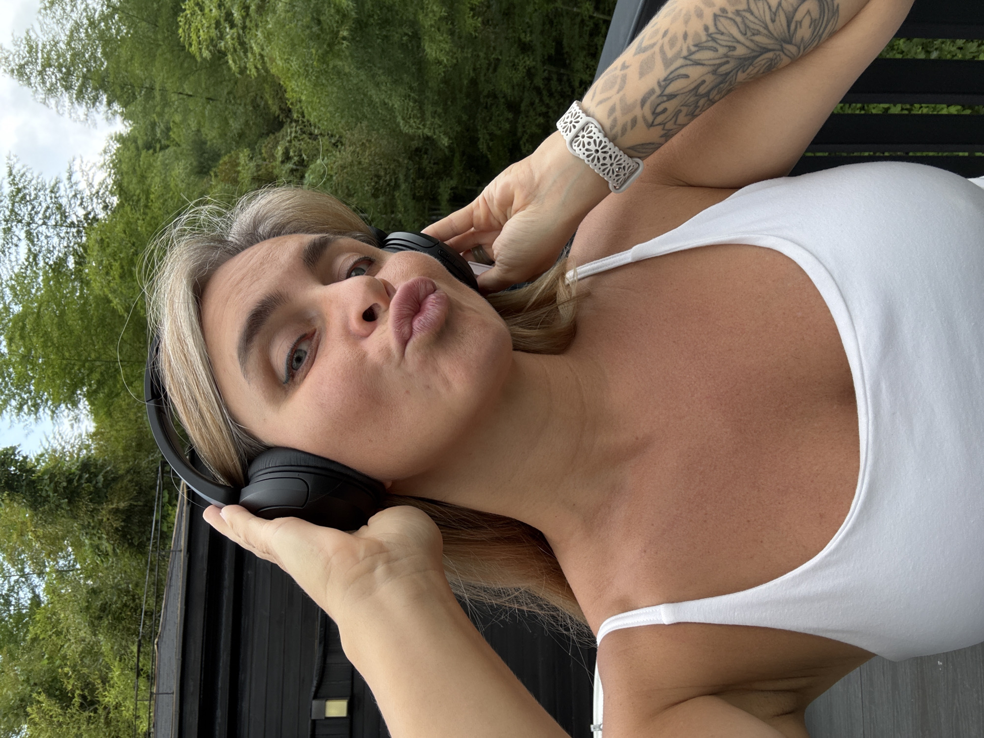 A medium shot, selfie-style image shows a woman with blonde hair and a white tank top. She is wearing black headphones and puckering her lips. She has a white bracelet on her right wrist, and a tattoo on her right arm. The background is lush green foliage and a dark building. The image appears to have been taken outdoors, on a balcony or deck. The context of Wuxing District, China, is not discernible from the visual information provided in the image.