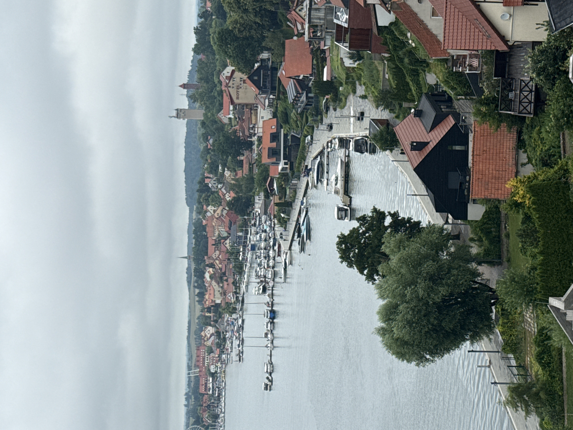 The image captures a view of Mikołajki, Poland, specifically the lakeside town and its marina. In the foreground, there are several modern houses with red tile roofs nestled into a lush, green hillside. A large, heart-shaped tree with green foliage dominates the left side of the foreground, with its trunk partially obscured by the hillside. Adjacent to the tree, a paved walkway runs along the water's edge, lined with a dark fence and lampposts.
The middle ground features a wide expanse of calm water, likely Lake Mikołajskie, filled with numerous docked boats of varying sizes, including sailboats and motorboats. A few of these boats are in motion or docked along piers. Along the waterfront, a dense collection of buildings, typical of a resort town, can be seen. These structures are a mix of older, traditional architecture with red roofs and more contemporary designs. The town extends up a gentle slope behind the marina.
In the background, the landscape is characterized by rolling hills covered in dense green trees. Several prominent structures stand out on the horizon: a tall, light-colored tower with a statue or figure on top, and a shorter, reddish-brown tower behind it, possibly a historical landmark or communication tower. The sky is overcast and a uniform light gray, suggesting a cloudy or possibly rainy day. The overall scene depicts a serene and scenic view of a lakeside town with a busy marina.