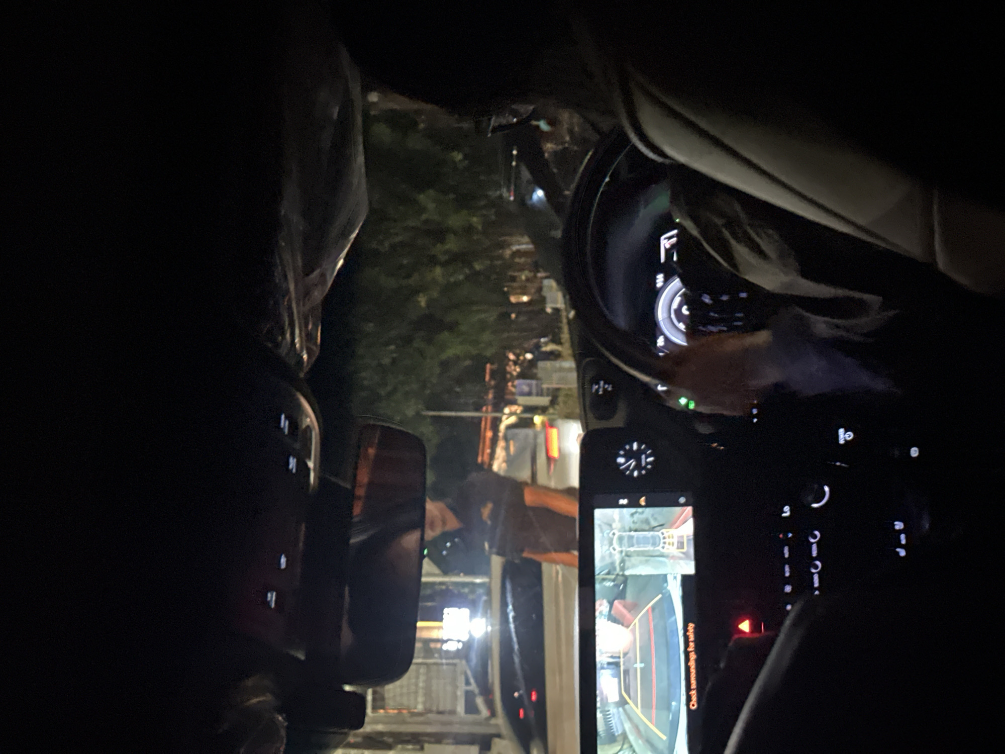 A nighttime scene inside a car, likely in South Jakarta, Indonesia, is depicted through the dashboard and rearview mirror. The car is positioned on a street lined with other vehicles and trees. A dashboard screen displaying a rearview camera feed is visible. The steering wheel and dashboard lights are also visible. The rearview mirror shows a reflection of a person standing behind the vehicle. The exterior environment is illuminated by street lights.