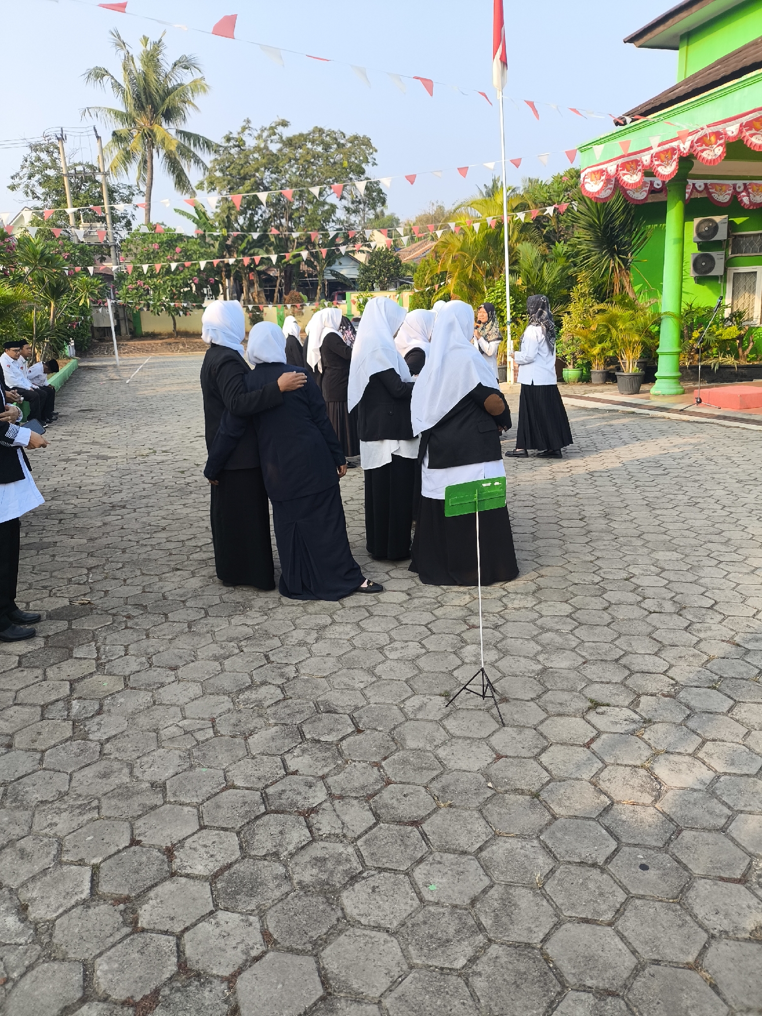 In Cilegon, Indonesia, a group of people, likely students, are gathered outdoors on a paved area with hexagonal stones. The scene appears to be at a school due to the presence of a school building with green paint and red and white decorative trim, along with a flagpole flying the Indonesian flag. Several individuals are wearing black outfits with white headscarves, suggesting a uniform or ceremonial attire. One person in the center appears to be speaking into a microphone, while others are looking towards them or interacting with each other. Some are standing, while others are seated on a green bench on the left side of the frame. Decorative red and white triangular flags are strung across the scene, common for national celebrations. The environment is bright and sunny, with trees and plants visible in the background.