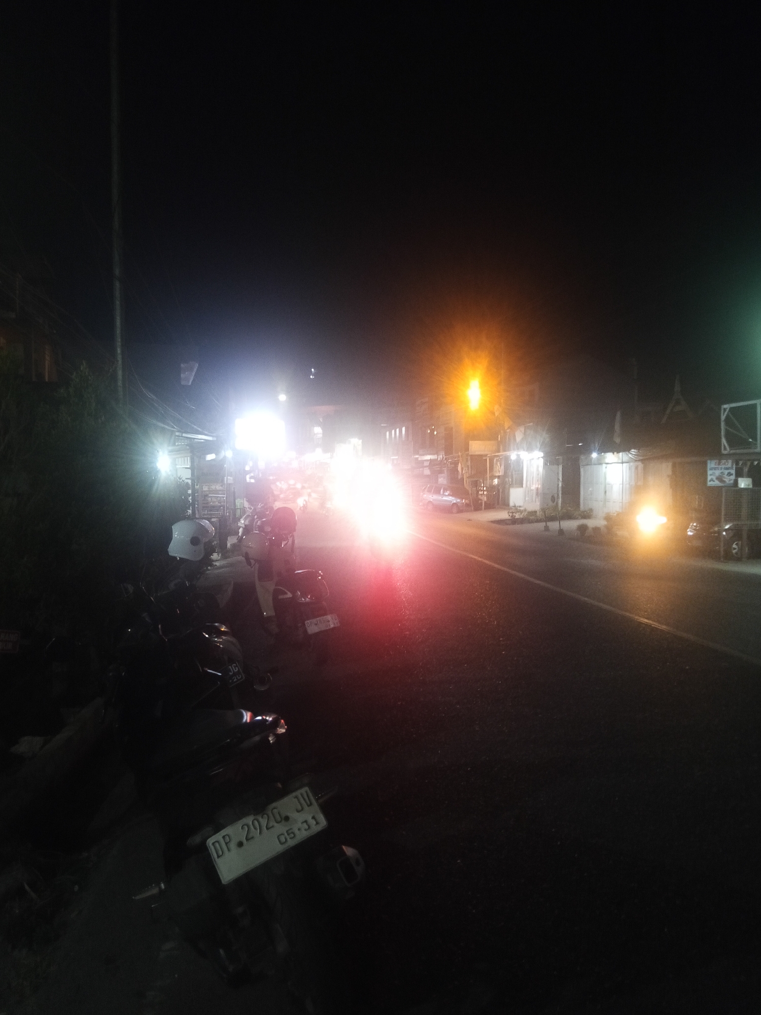 The image depicts a nighttime street scene in Makale, Indonesia, characterized by artificial lighting and the presence of vehicles. In the foreground, several motorcycles are parked, one of which has a license plate reading "DP. 2920. JV 05-31." The road is visible, with streetlights and building lights illuminating the area. There is a significant amount of light glare, particularly from headlights and streetlights, creating a hazy effect. A few buildings with shopfronts line the street. The overall impression is of a busy urban environment at night.