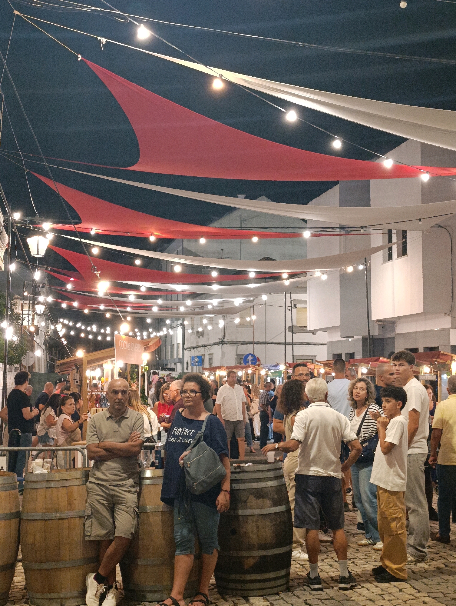 This is a vertical, eye-level shot capturing a nighttime street scene at a festival or market in Palmela, Portugal. The foreground is dominated by three large wooden barrels, with a man in a light brown polo shirt and cargo shorts leaning against the leftmost barrel, arms crossed, and a woman in a dark blue t-shirt and shorts standing next to the middle barrel. The woman has a backpack slung over her shoulder. Behind them, a crowd of people is milling about, with various stalls and vendor booths visible further down the street. String lights crisscross overhead, illuminating the scene, and large red and white fabric awnings are draped between buildings, creating a festive canopy. The buildings appear to be white or light-colored with dark windows. The ground is paved with cobblestones. The overall atmosphere is lively and social.