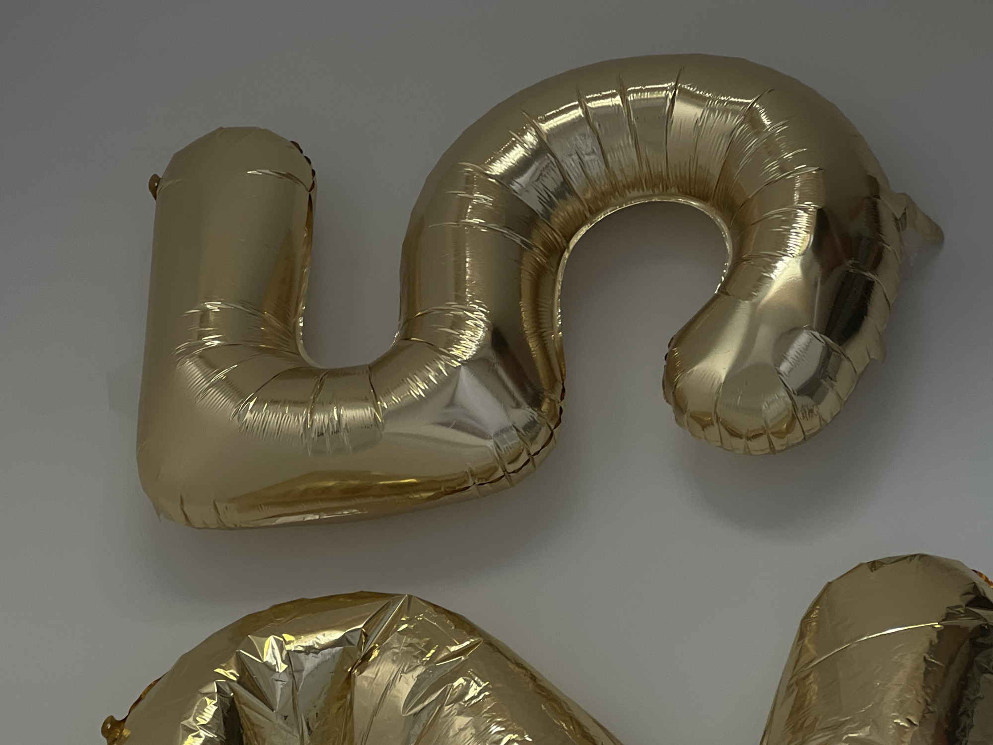 The image shows two large, gold Mylar balloons that form the numbers "2" and "5". The balloons are inflated and appear to be attached to a white wall. The lighting suggests an indoor environment. The balloons are positioned to the left and center of the frame, with the number "2" partially visible on the left and the number "5" prominently displayed in the center. There are no people or other objects visible in the image. The location is not specified in the image.
