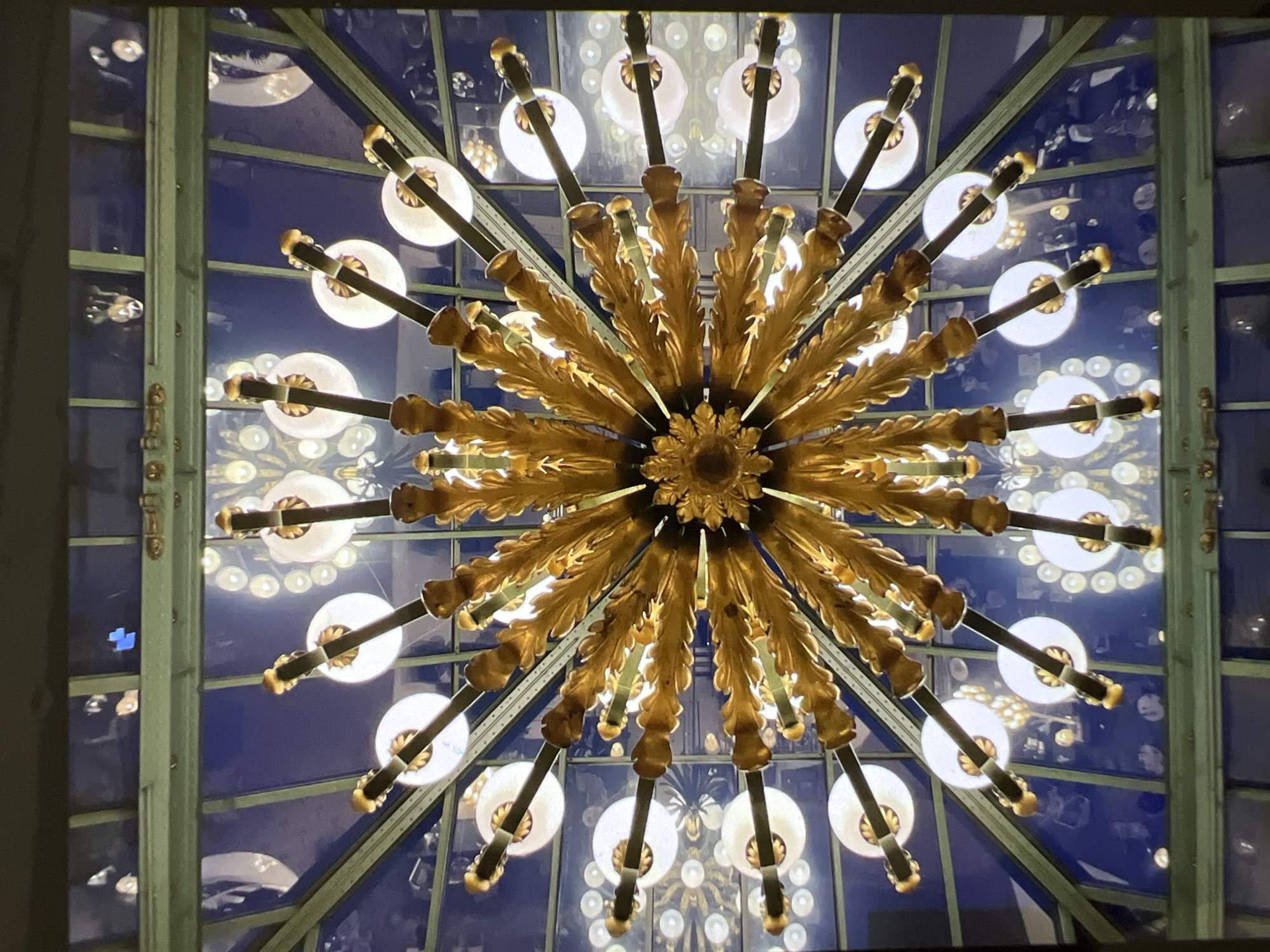 An ornate, golden chandelier with starburst-like arms and leaf-shaped accents hangs in the center of the frame, suspended from a glass ceiling with a dark blue background. The chandelier has multiple lights, each with a white circular shade and a visible filament. The ceiling is framed by green metal beams and glass panes, revealing reflections of the chandelier and other lights, as well as some indistinct shapes. The perspective is from below, looking directly up at the chandelier and the ceiling. The scene appears to be indoors, likely in a grand hall or conservatory. The image has a high contrast and a shallow depth of field, with the chandelier in sharp focus and the background elements appearing softer. There are no people visible in the image.