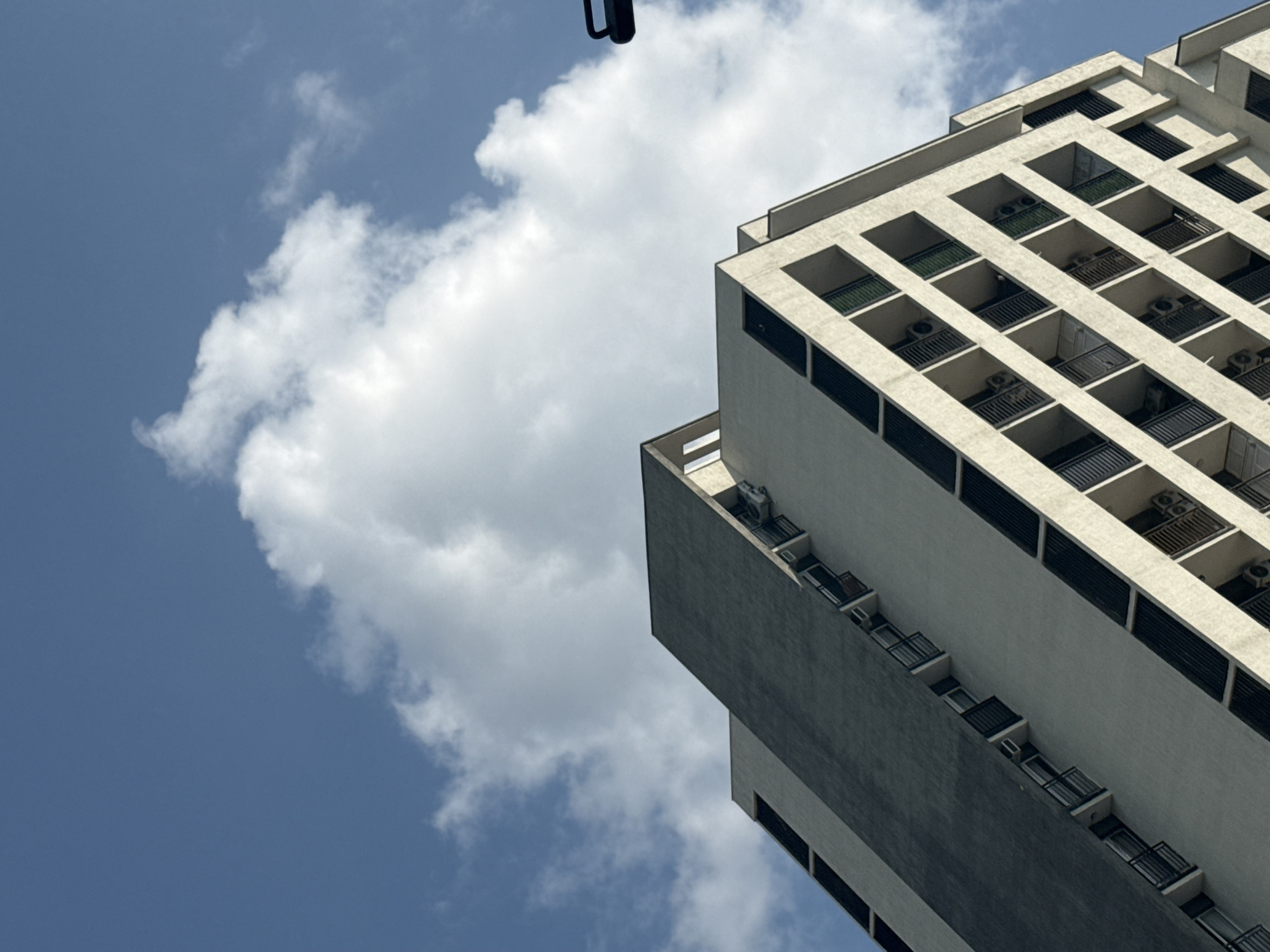 The image shows the upper portion of a modern apartment building in Tbilisi, Georgia, viewed from a low angle against a blue sky with scattered white clouds. The building features a distinctive architectural design with a dark, sloped facade on the left and a lighter, stepped facade on the right. The lighter facade has multiple balconies with black railings, and air conditioning units are visible on some of them. The image is a still photograph taken outdoors during the daytime.