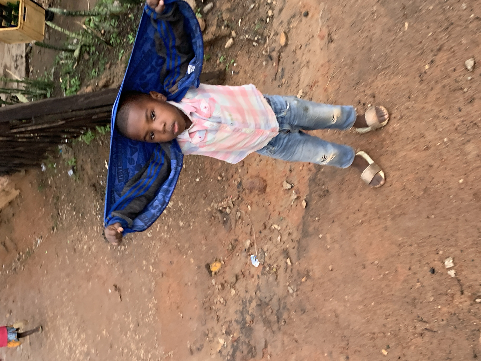 The image shows a young boy standing on a dirt path in what appears to be an urban or semi-urban environment in Jos, Nigeria. The boy is holding a blue blanket or garment over his head, partially obscuring his face. He is wearing a light pink and white checkered shirt and ripped blue jeans. On his feet, he has light-colored slide sandals.
The background consists of a dirt ground with scattered debris. To the left, another child, wearing a pink top and yellow shorts, is partially visible, walking away from the camera. In the middle ground, there is a wooden fence and some plants. To the right, a yellow crate filled with green bottles, possibly beverages, is visible, suggesting a commercial or informal market area. The overall lighting suggests it may be daytime, possibly overcast or late afternoon. The scene depicts a common sight in many parts of Jos, Nigeria, with its unpaved roads and children playing outdoors.