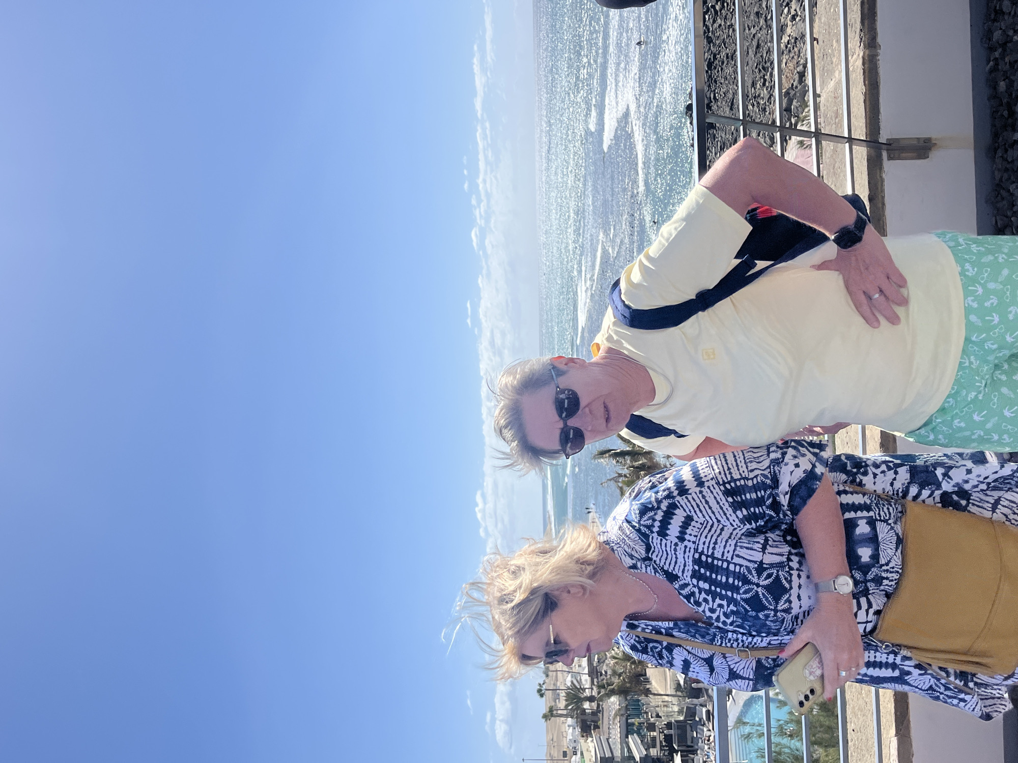 Two middle-aged people are posing for a picture, with the man in the back wearing a yellow shirt and green shorts, while the woman in front is wearing a blue and white patterned shirt and has a tan purse. They are on a platform overlooking the sea and possibly a city in Maspalomas, Spain. The sky is blue with few clouds, and the sea is wavy. The woman holds a smartphone.