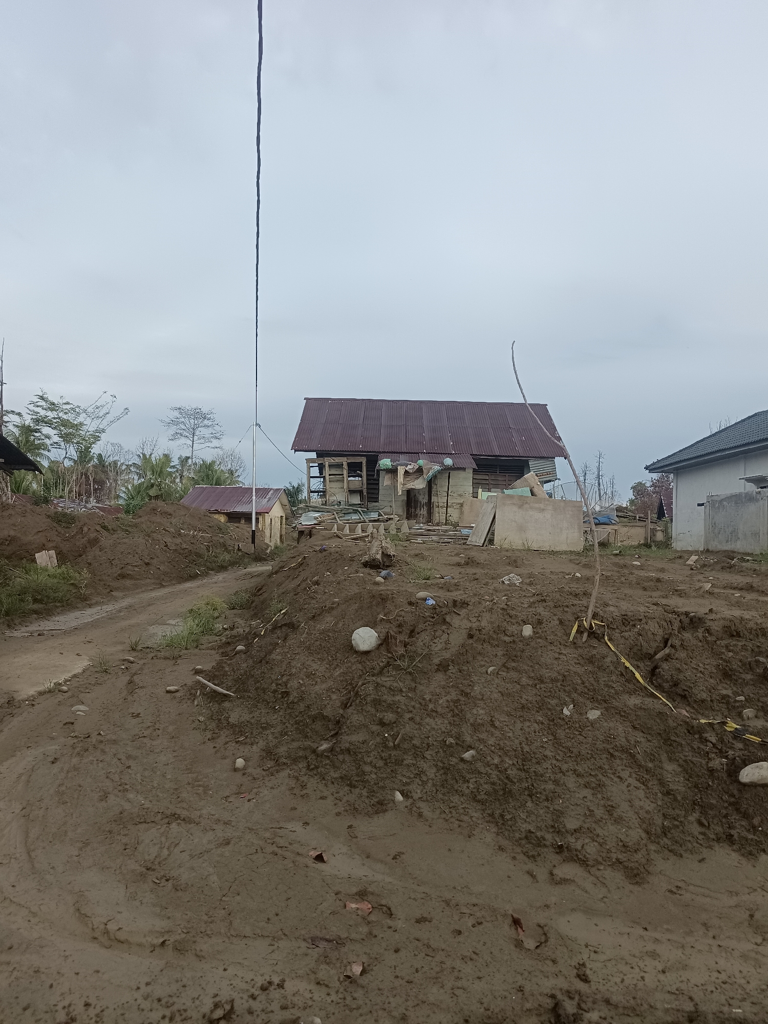 The image depicts a scene in Meunasah Raya, Indonesia, characterized by the aftermath of what appears to be a natural disaster, likely a landslide or flood, given the significant accumulation of mud and debris. In the foreground, a muddy path winds through large mounds of dark soil. Several small rocks and some sparse vegetation are scattered across these mounds.
Further into the image, several buildings are visible. One prominent structure has a corrugated metal roof and wooden walls that appear damaged or unfinished, with scaffolding-like elements. Adjacent to it, a smaller building with a similar metal roof stands. To the right, a more modern, lighter-colored building with a dark roof is partially visible. In the background, trees and other vegetation are present, some of which appear to have been affected by the disaster. A tall, thin pole, possibly a utility pole, extends vertically into the overcast sky. The overall atmosphere is somber, with a neutral, cloudy sky. There are no people visible in the image. The scene suggests a community in a state of disarray and potential rebuilding efforts.