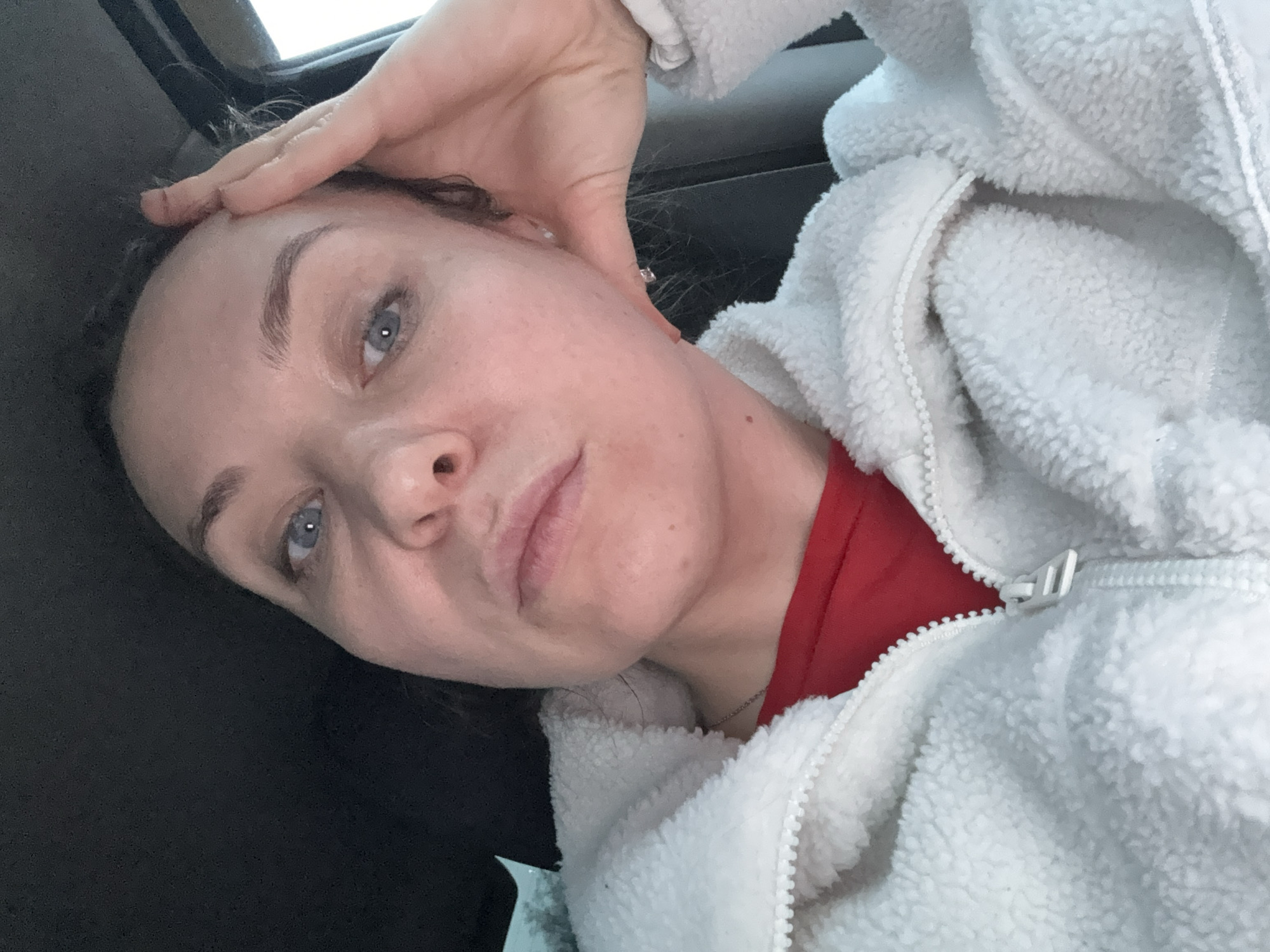 A young woman is captured in a close-up, angled selfie, likely taken indoors or within a vehicle in Chaikovsky, Russia. She has light blue or grey eyes and dark eyebrows. Her dark hair appears pulled back, with a few wisps visible around her temples. Her right hand rests gently on her forehead.
She is dressed in a white, textured, zippered jacket, possibly made of sherpa or fleece material, with a vibrant red shirt visible underneath. A small, shiny stud earring is visible in her right ear (viewer's left), and a thin, silver-colored chain necklace can be seen peeking from beneath her jacket near her neck. The warm clothing suggests a cool or cold environment, consistent with the climate in Chaikovsky, Russia.
The background is dark and indistinct, potentially hinting at a car interior or a dimly lit room, with a soft, diffused light source visible at the top edge of the frame. Her expression is neutral and calm as she looks directly towards the camera. There is no visible text in the image.