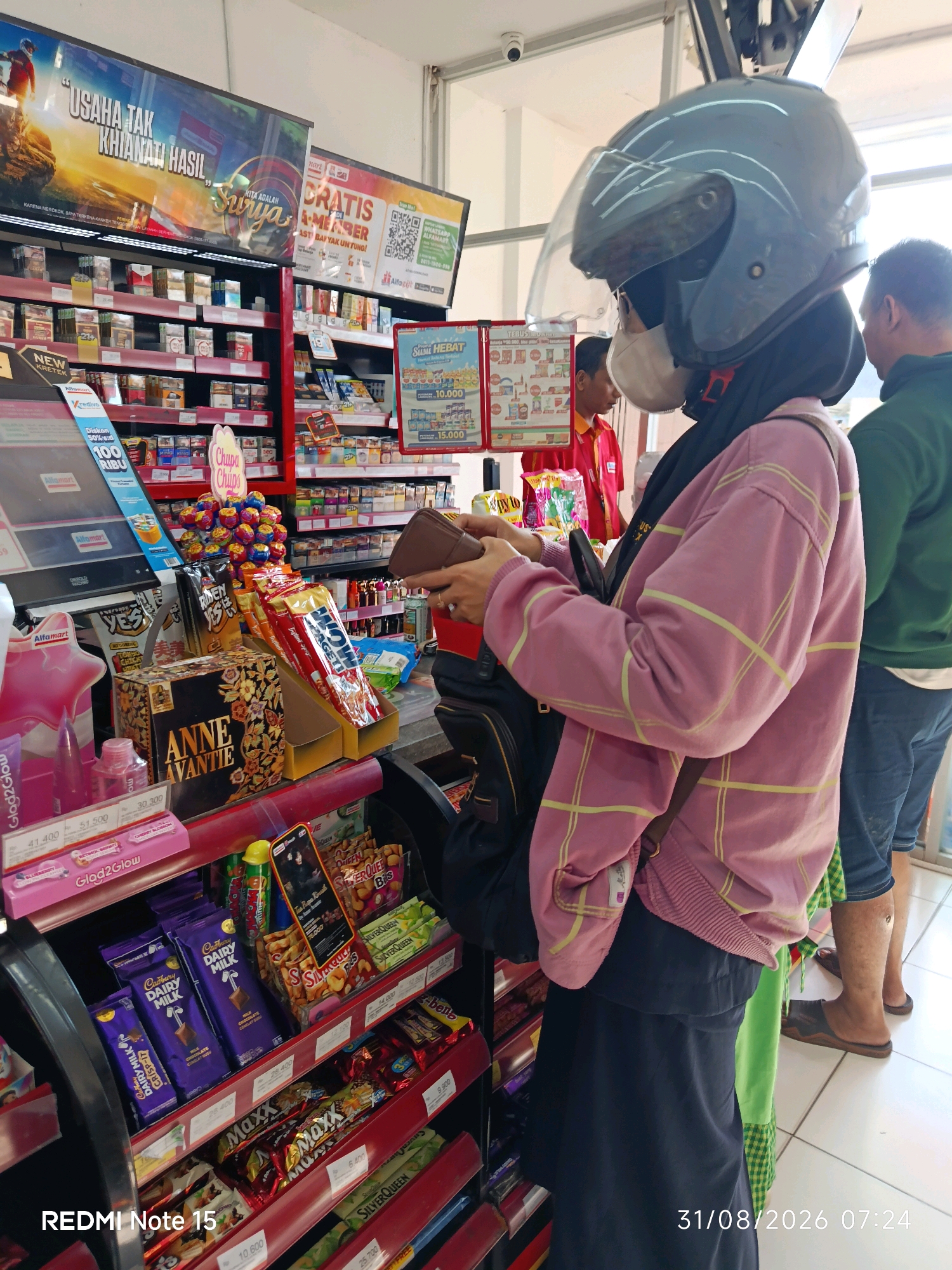 The image depicts the interior of a minimarket, possibly an Alfamart, in Keboansikep, Indonesia. A person wearing a grey helmet, a face mask, a pink plaid long-sleeved shirt, and a dark skirt stands at the checkout counter, holding a brown wallet. Another person in a red uniform, likely an employee, is visible behind the counter. To the right, a person in a green shirt and blue jeans is waiting. The shelves are stocked with various products, including cigarettes, snacks, and chocolates. A promotional display for "Chupa Chips" is prominently featured. The lighting suggests an indoor environment with artificial lights. The date and time stamp "31/08/2026 07:24" is visible in the bottom right corner.