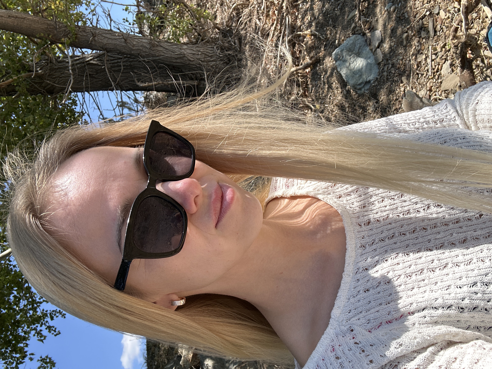 A blonde woman wearing sunglasses and a white knitted sweater is outdoors. She has light-colored hair that is long and straight, and she is wearing a hoop earring in her left ear. The background shows a tree with rough bark and branches, and the ground is covered in rocks and dry vegetation. The sky is blue, and there are some hints of foliage from trees. The scene appears to be a natural outdoor setting, possibly a park or a wooded area. The image doesn't provide specific clues about its location within the city of Gornyi Erik, Russia, as it depicts a generic natural environment.