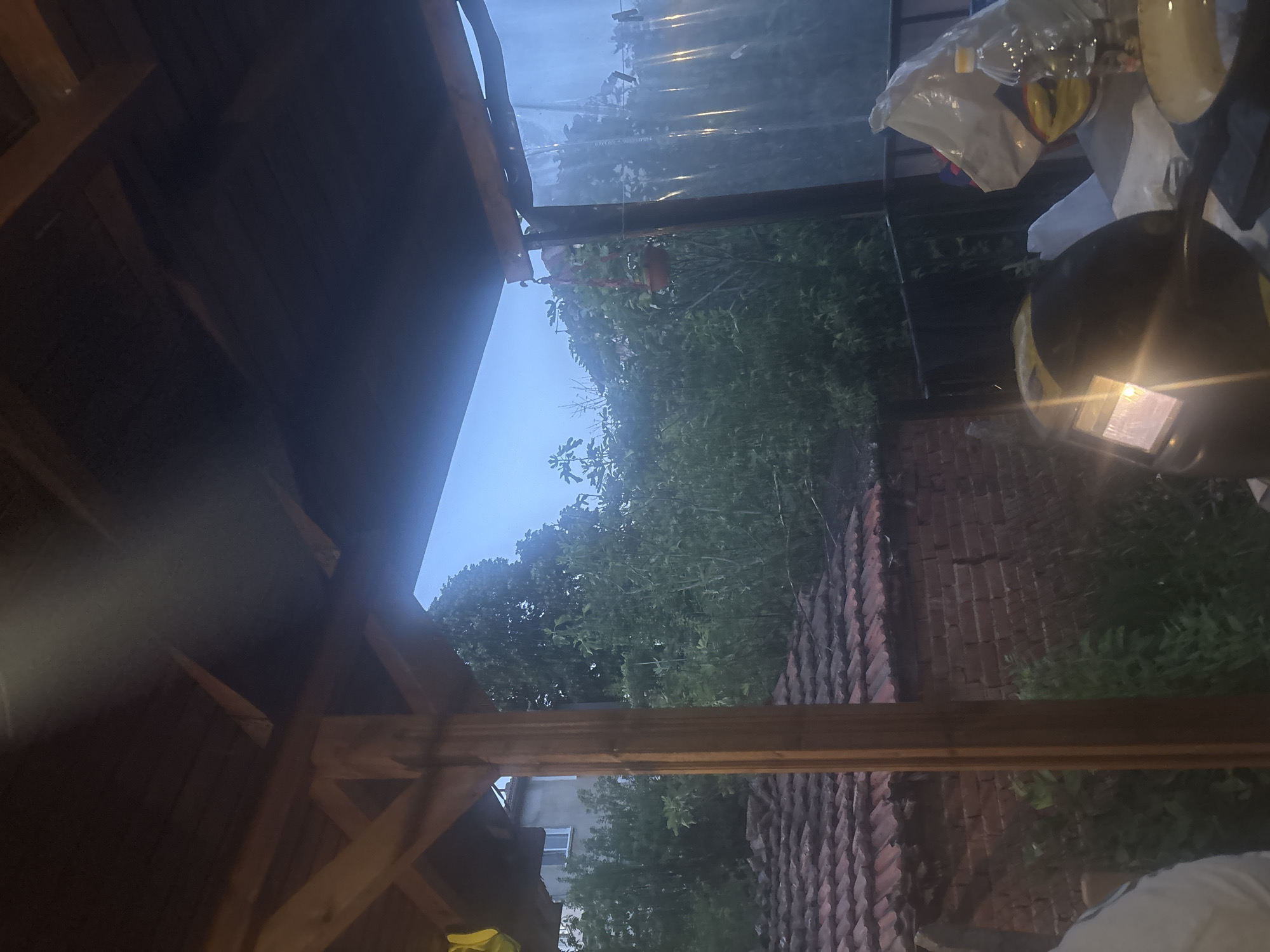 The image depicts an outdoor scene in Varshilo, Bulgaria, viewed from underneath a wooden pergola or shelter during dusk or an overcast period. The overhead structure comprises dark wooden ceiling planks supported by lighter-toned wooden beams and posts.
In the foreground, on the right, a dark welding helmet with an illuminated rectangular visor rests on a surface, likely a table. Adjacent to it are a white plastic bag, possibly a grocery bag with a yellow and red logo, and a transparent plastic bottle containing liquid. The edge of a light-colored frying pan is also partially visible on the table.
Beyond the shelter, the environment features residential structures and dense vegetation. On the left, a section of a reddish-brown tiled roof is visible above a red brick wall. Behind these, extensive green foliage, including bushes and trees, fills the midground. A light-colored building with white windows is partially discernible through the trees on the far left. A prominent vertical wooden post of the pergola frames the left side of this exterior view.
On the right side of the exterior, a small red potted plant hangs from a wooden beam. To its right, a translucent plastic sheet, serving as a curtain or barrier, hangs vertically, attached by string and clothespins. The sky in the background is a muted blue, consistent with twilight or an overcast condition. No individuals are explicitly visible, though a partial white garment in the bottom left suggests a person's presence just outside the frame.