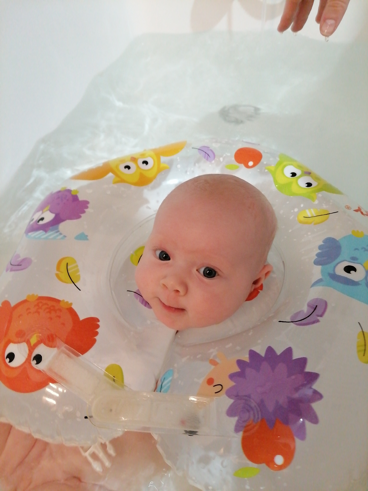 A baby is floating in a bathtub with a neck floatation device. The float is white with various cartoon owls and other illustrations. The baby is looking at the camera with a slight smile. The baby is surrounded by water. A hand is visible above the water, possibly belonging to a caregiver. The location of Trubacheyevka, Russia, is not discernible from the image.