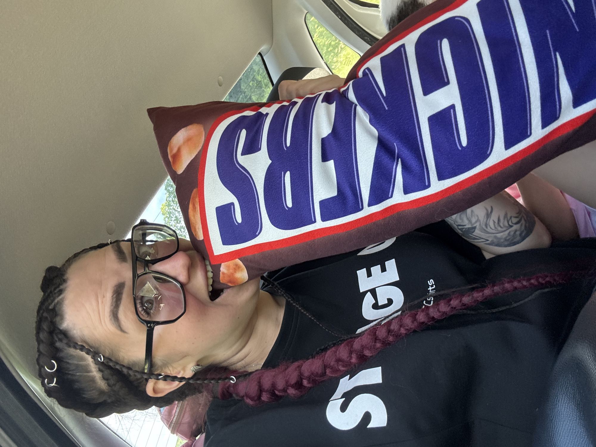 An adult Caucasian woman with dark hair styled in braids, some adorned with silver rings, wears large black-framed glasses and a black t-shirt with white text. She is in the passenger seat of a vehicle, her head tilted to the side as she bites into a large pillow shaped like a Snickers candy bar. The pillow has the brand name "SNICKERS" in large, bold letters, with the "S" partially obscured. Her mouth is open, revealing her teeth and tongue against the pillow. A portion of a tattoo is visible on her right arm. The interior of the car is visible, including the dashboard, windshield, and the car's headliner. Outside the vehicle, green foliage and trees suggest a daytime setting, possibly a suburban or rural area. The lighting indicates it is daytime.