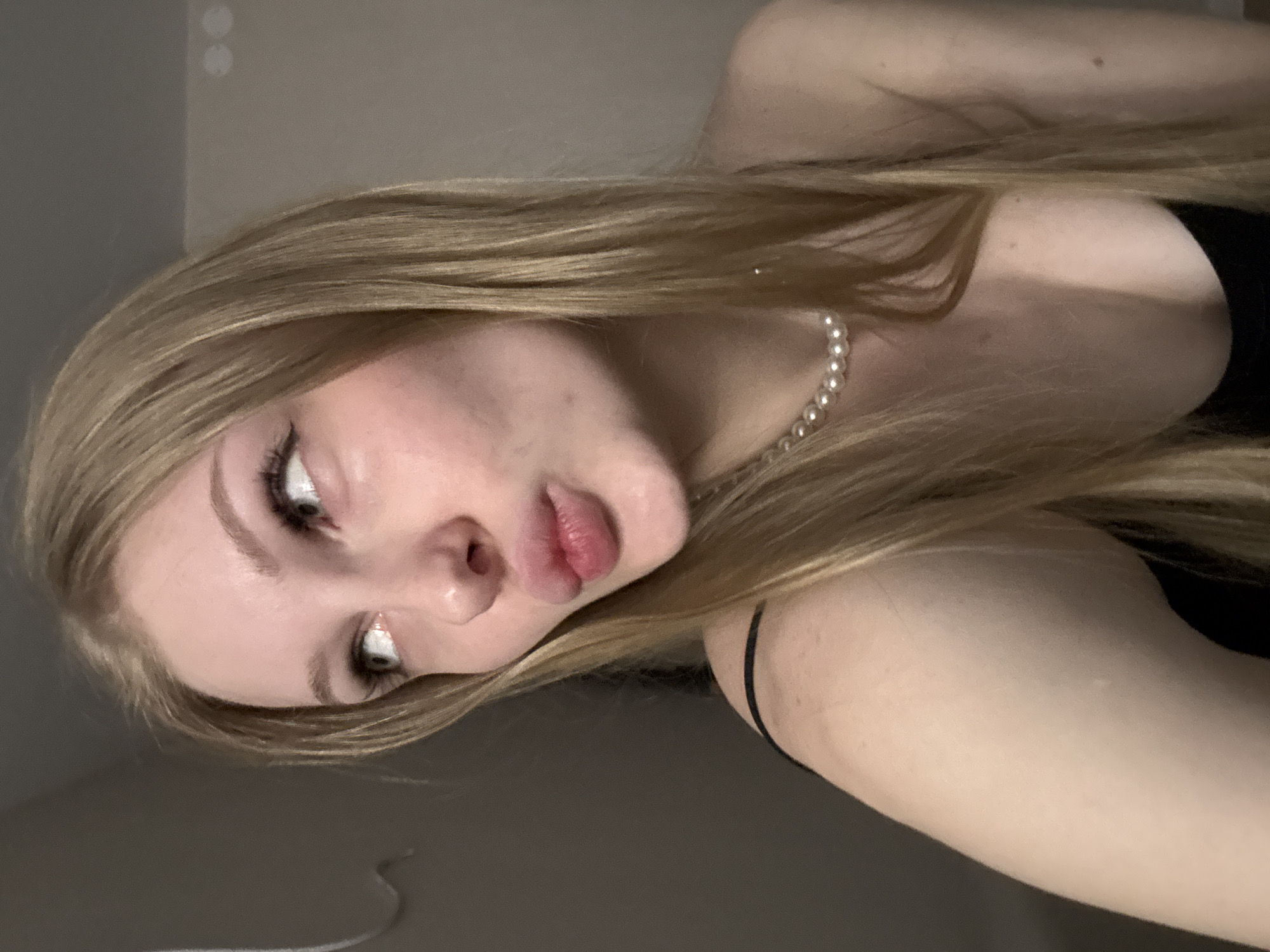 The image shows a close-up portrait of a young Caucasian woman with blonde hair and fair skin, looking to her left. She has her lips puckered as if for a kiss. She is wearing a black top and a pearl necklace. The background is a plain, neutral-colored wall. There is no indication of the location in the city of Perm, Russia.