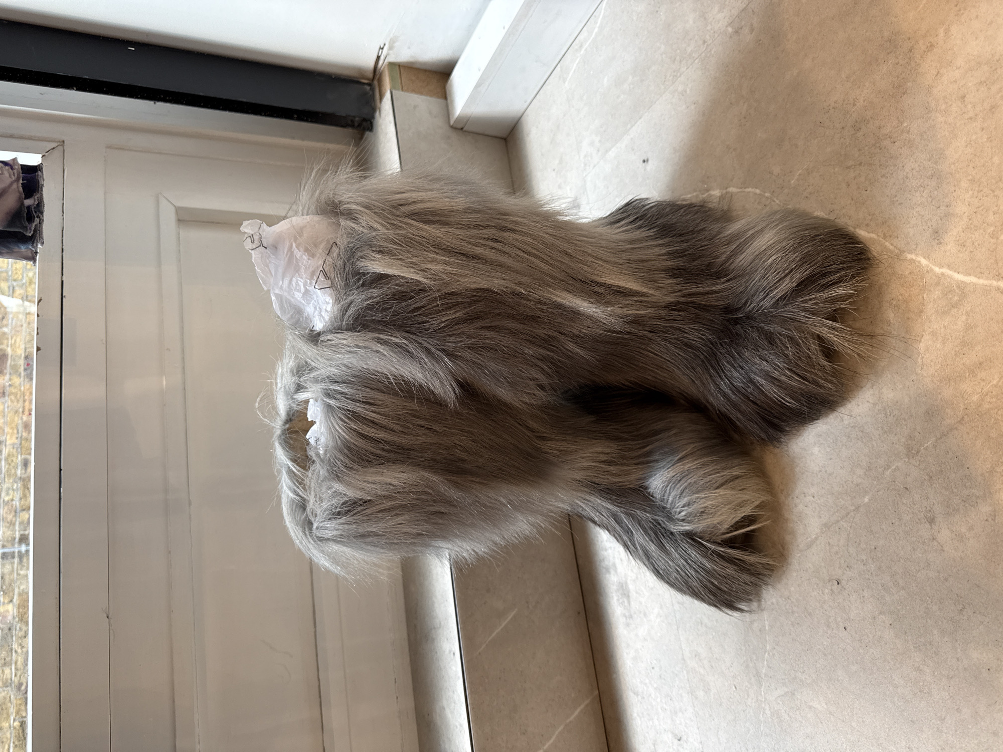 The image shows a pair of grey, furry boots resting on a light-colored tiled floor. The boots are partially wrapped in white tissue paper at the top. A white door with a black frame is visible on the left side of the image. The setting is indoors. It is not possible to determine if this image was taken in the city of London or more generally, the United Kingdom.