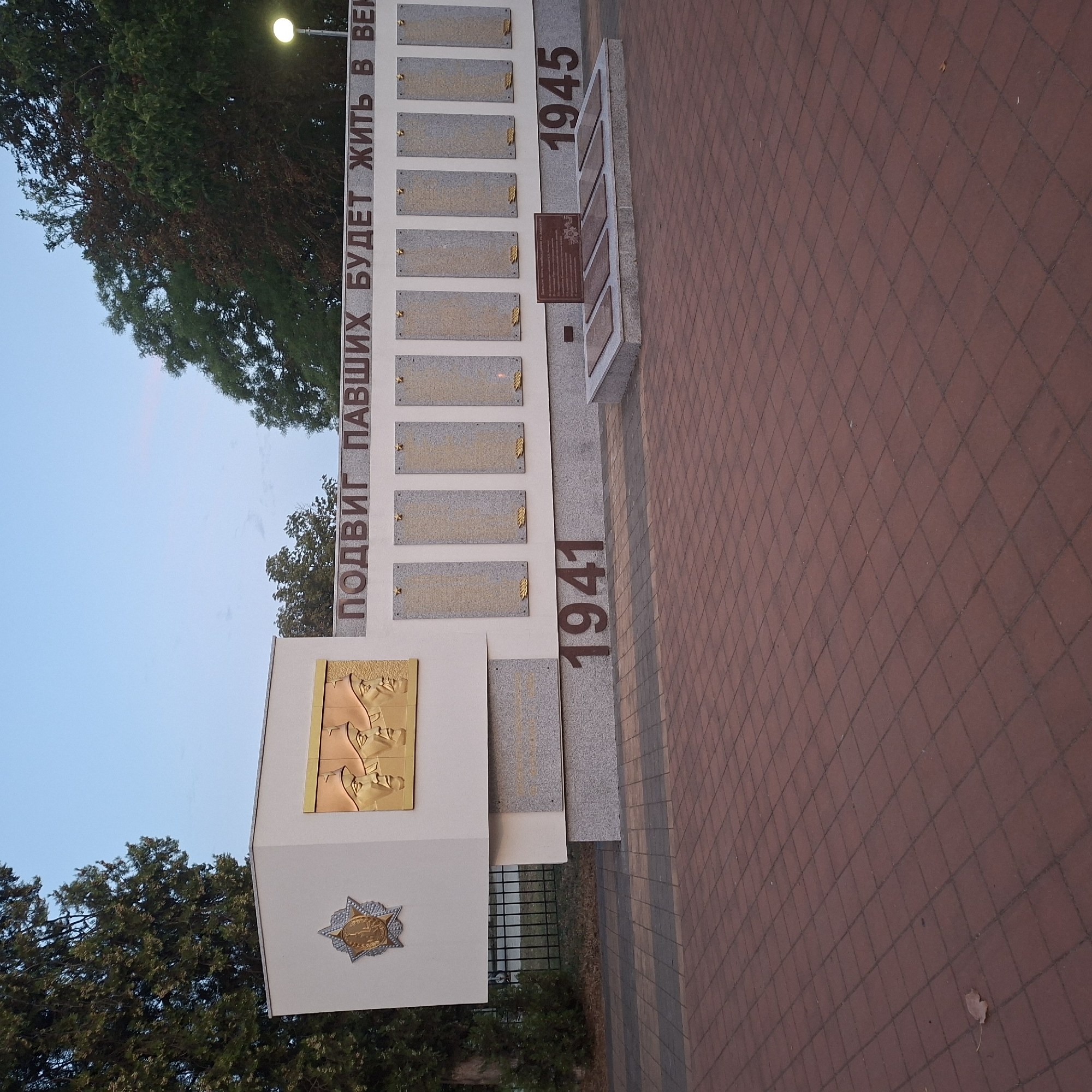 The image depicts a monument with memorial plaques, likely dedicated to World War II. The monument features a large plaque with three profiles of soldiers and a star emblem. Below this, a row of rectangular plaques with inscriptions is visible, along with the numbers "1941" and "1945". A lower section of the monument has several flat memorial stones arranged in a semi-circle. The monument is set against a backdrop of trees and a clear sky. The ground in front is paved with reddish-brown square tiles, and a single fallen leaf is visible on the paving. The location is identified as Anapskaya, Russia.