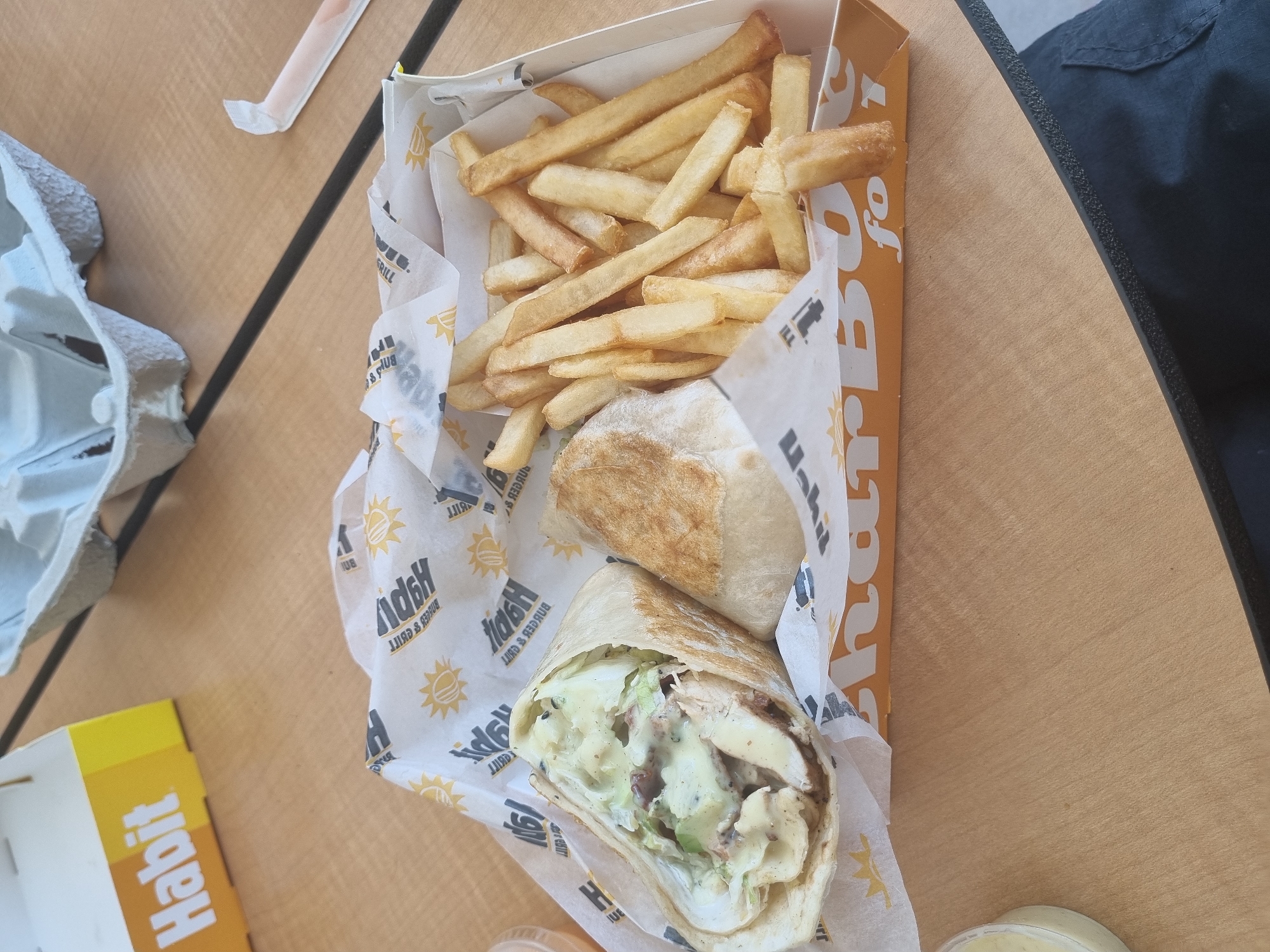 A close-up, eye-level shot shows a meal from Habit Burger Grill, served on a wooden table. The meal consists of a wrap with chicken and sauce, and a side of french fries. The food is presented in a white paper-lined tray with the Habit Burger Grill logo. A yellow Habit Burger Grill box is visible in the background. A grey cardboard egg carton rests on the table behind the meal. The setting is likely indoors, possibly a restaurant or a food court. No people are visible in the image. The overall impression is a casual meal setting. The image does not provide any information about the location within the city of Vancouver, United States of America.