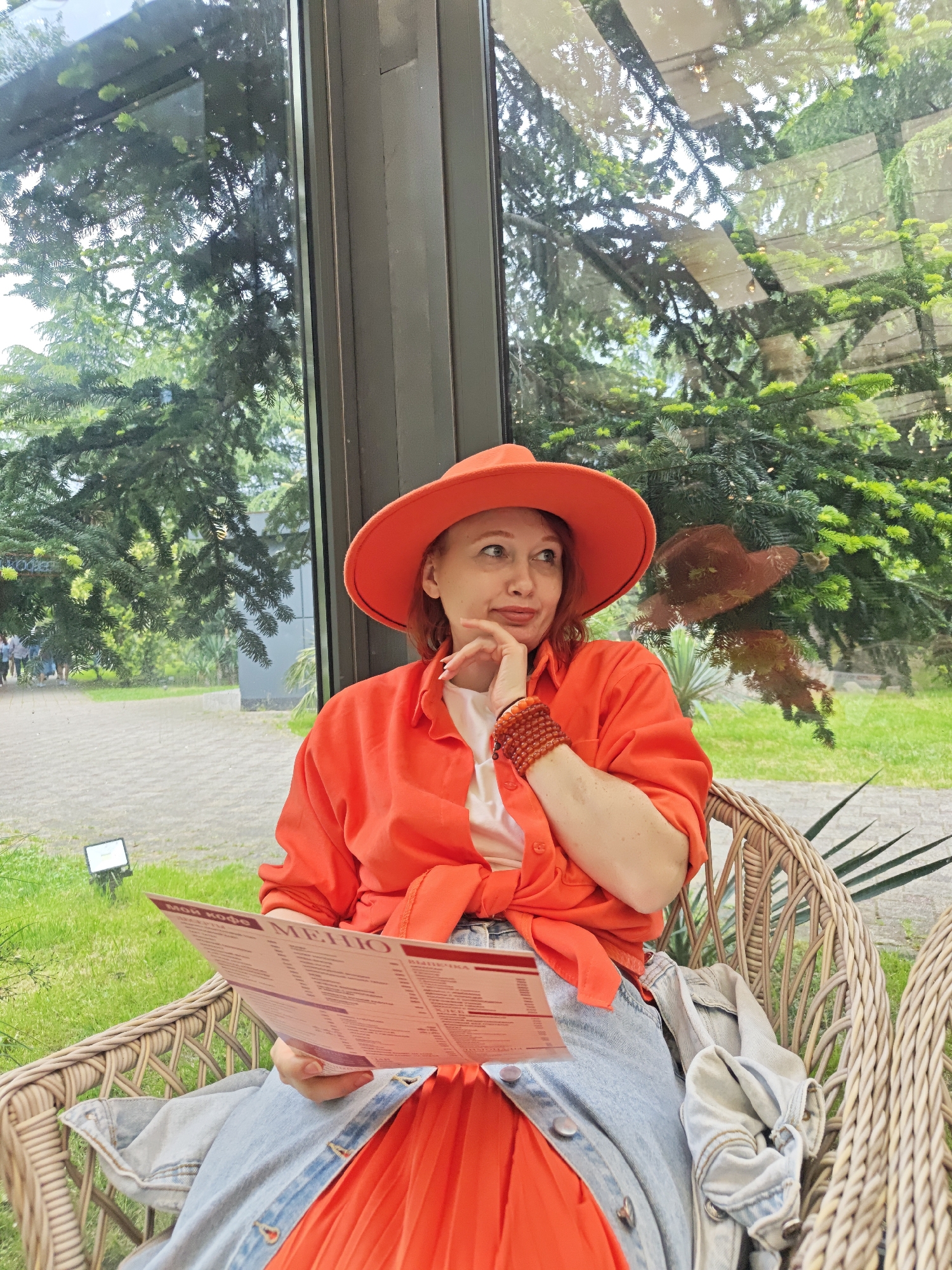The image shows a woman sitting in a wicker chair, possibly in a restaurant or cafe, given the menu she's holding. She's wearing an orange hat, a matching orange shirt tied at the waist, a white top underneath, a light blue jean jacket, and a pleated orange skirt. She has a reddish-ginger hair color. She is wearing beaded bracelets on her wrist and has her hand to her face in a pensive pose, looking upwards. A large window behind her shows trees and a path or patio outside. Based on the reflections in the window and the style of architecture and foliage, it is likely that the location is in the city of Sochi, Russia.