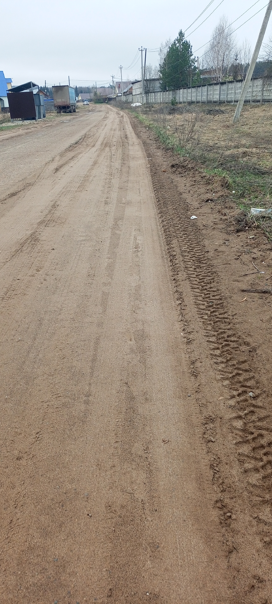 The image depicts a straight, unpaved dirt road stretching into the distance under an overcast sky. The road surface is a light brown, sandy earth, heavily marked with numerous tire tracks. To the immediate right of the road, the ground transitions to rougher terrain with sparse, dry vegetation and more pronounced tire treads running parallel to the road.
In the mid-ground and background, various structures line both sides of the road. On the left, there are what appear to be sheds or storage containers, including a dark brown one and a lighter metallic blue one, next to a building with a blue roof. Further down, a large, older truck with a box-like cargo area is parked. A white car is visible further down the road.
On the right side of the road, a long, tall fence constructed from grey concrete panels stretches into the distance, separating the road from an area with more mature trees, including evergreens and bare deciduous trees, suggesting a cooler season or early spring. Utility poles with overhead wires run alongside the road on both sides. The overall setting suggests a rural or semi-rural area, possibly a village or a developing residential zone. There is no visible activity or specific event taking place, and no discernible text. The light suggests it is daytime, likely an overcast day.