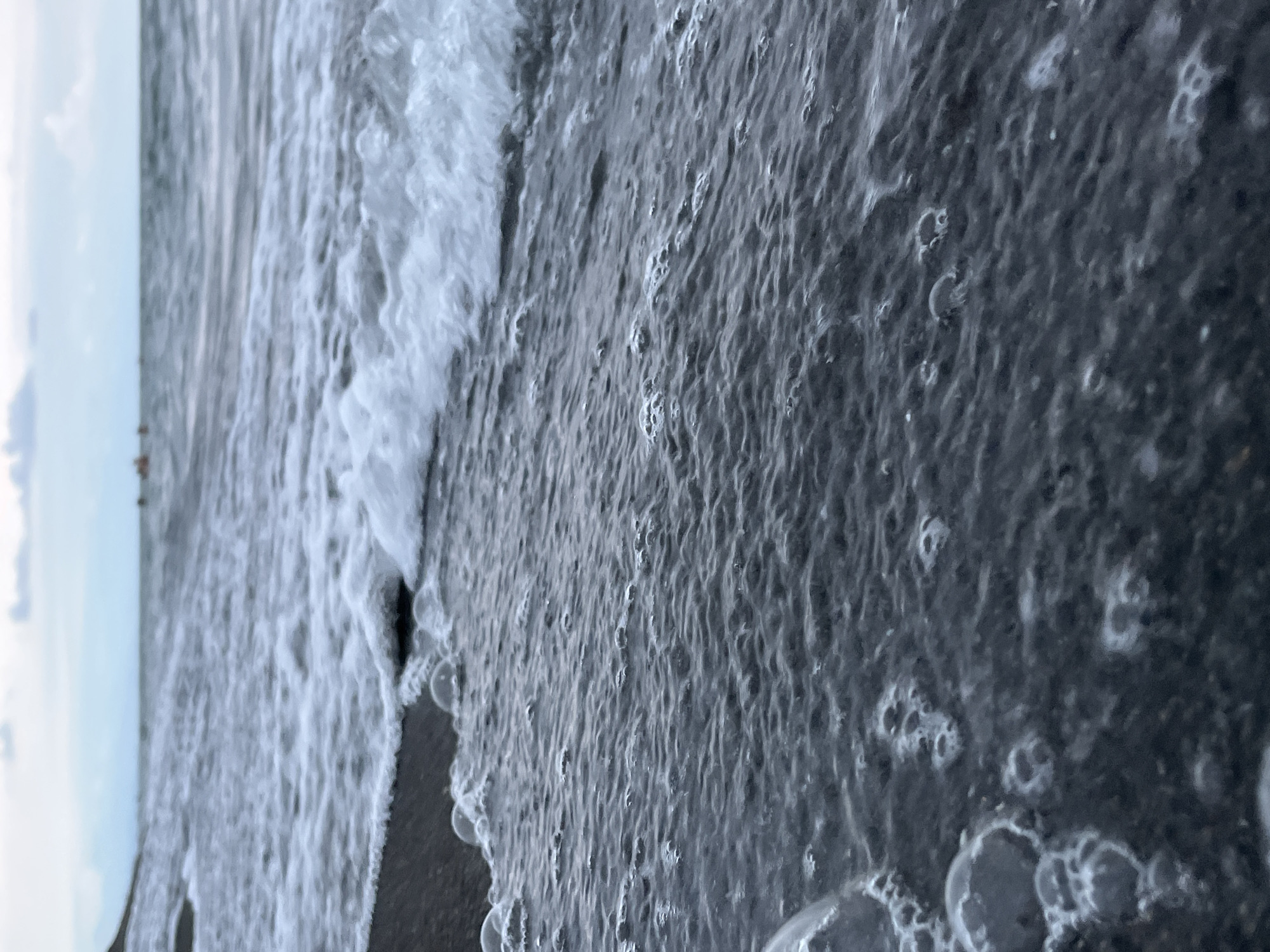 The image shows a close-up view of the sea and a black sand beach in Ureki, Georgia. The foreground is dominated by dark, wet sand with many small bubbles and foam from the waves. The water in the mid-ground is choppy with white foam. In the background, the sea stretches out to the horizon under a cloudy sky. A few indistinct figures can be seen in the distance on the water, possibly people on boats or swimming. The overall impression is of a slightly overcast day at the coast.