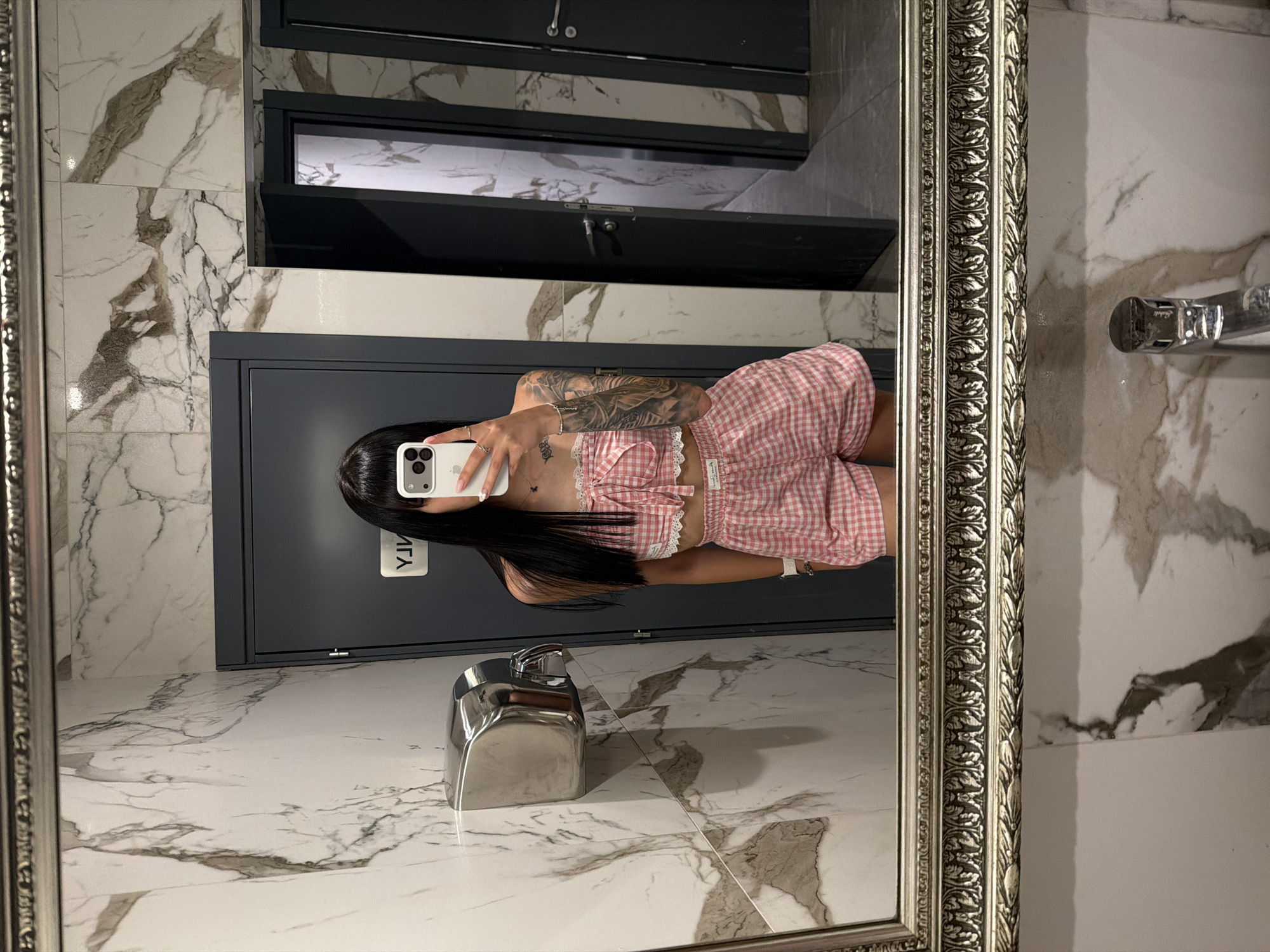 The image shows a young woman with long dark hair taking a mirror selfie in a public restroom. She is wearing a pink and white gingham two-piece set consisting of a cropped top with lace trim and matching shorts. Her left arm and shoulder are visible, revealing extensive tattoos. She is holding a smartphone in her right hand, which is angled to capture her reflection. The restroom features marble-patterned tiles on the walls, a silver hand dryer mounted on the left wall, and dark grey doors, one of which appears to be for a stall. The floor is also visible, with a marble-like pattern. The mirror is ornate and framed with a decorative silver border. The overall impression is a stylish, modern public restroom. The location is Plovdiv, Bulgaria.
