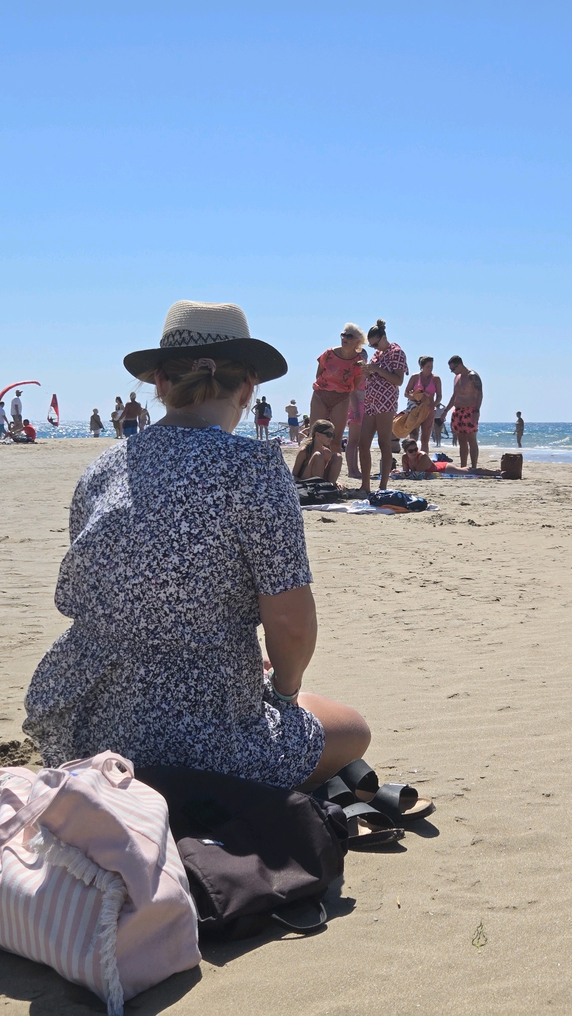 The image depicts a sunny day at Prasonisi beach in Greece, characterized by a clear blue sky and a sandy shoreline. In the foreground, a woman wearing a patterned dress and a wide-brimmed hat sits facing away from the camera, with a pink and white striped beach bag and a black backpack placed beside her. Black sandals are also visible.
In the midground and background, several groups of people are engaged in various beach activities. Some individuals are standing and interacting, while others are sunbathing or wading in the ocean. The presence of a red kite suggests activities like kitesurfing may be taking place further out on the water. The overall scene conveys a relaxed atmosphere typical of a summer beach day.