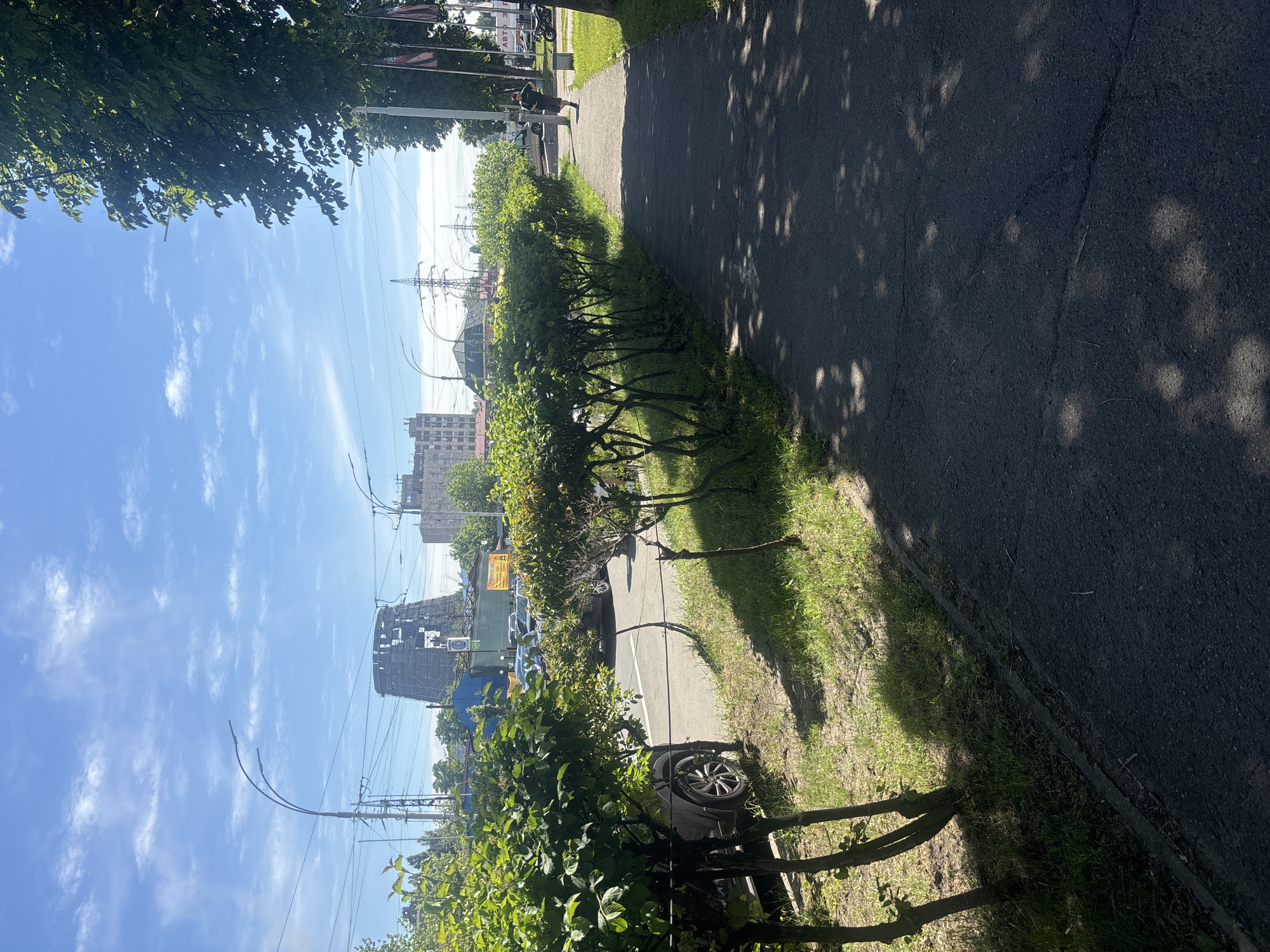 The image depicts a sidewalk in Kyiv, Ukraine, on a sunny day. The sidewalk runs along a street lined with trimmed green bushes on the left and a tree on the right. Beyond the bushes, cars are parked, and a distinctive, partially constructed cooling tower with scaffolding is visible in the background. Several utility poles with overhead wires and streetlights are also present. Further in the distance, apartment buildings and other structures indicate an urban environment. A person is seen jogging on the sidewalk in the mid-ground, and another person is standing near a parked motorcycle on the far right. The ground on the sidewalk is asphalt, with shadows of tree leaves cast upon it. The sky is blue with scattered white clouds.